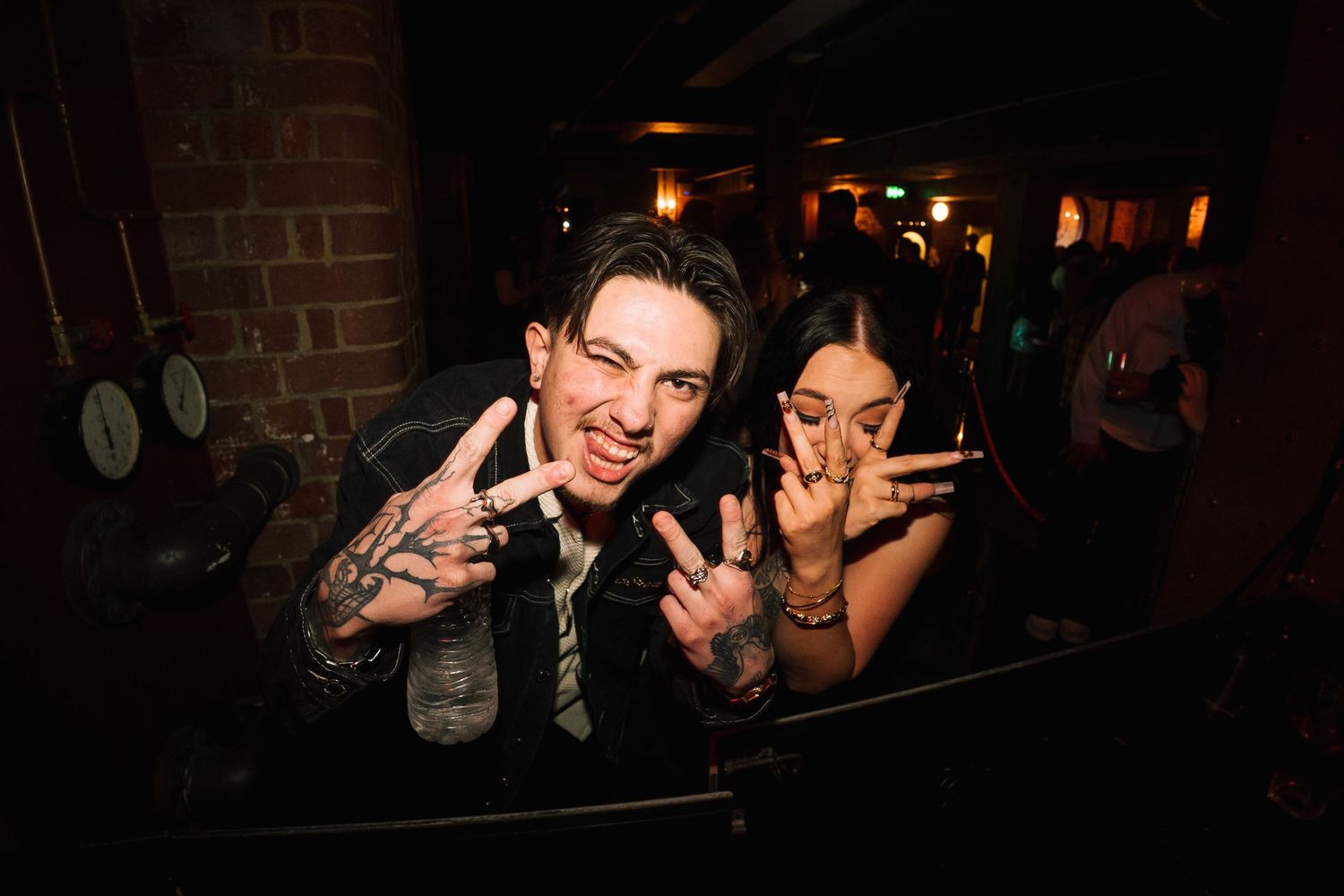 Two people making peace signs with their hands while posing together at a dimly lit bar or nightclub.