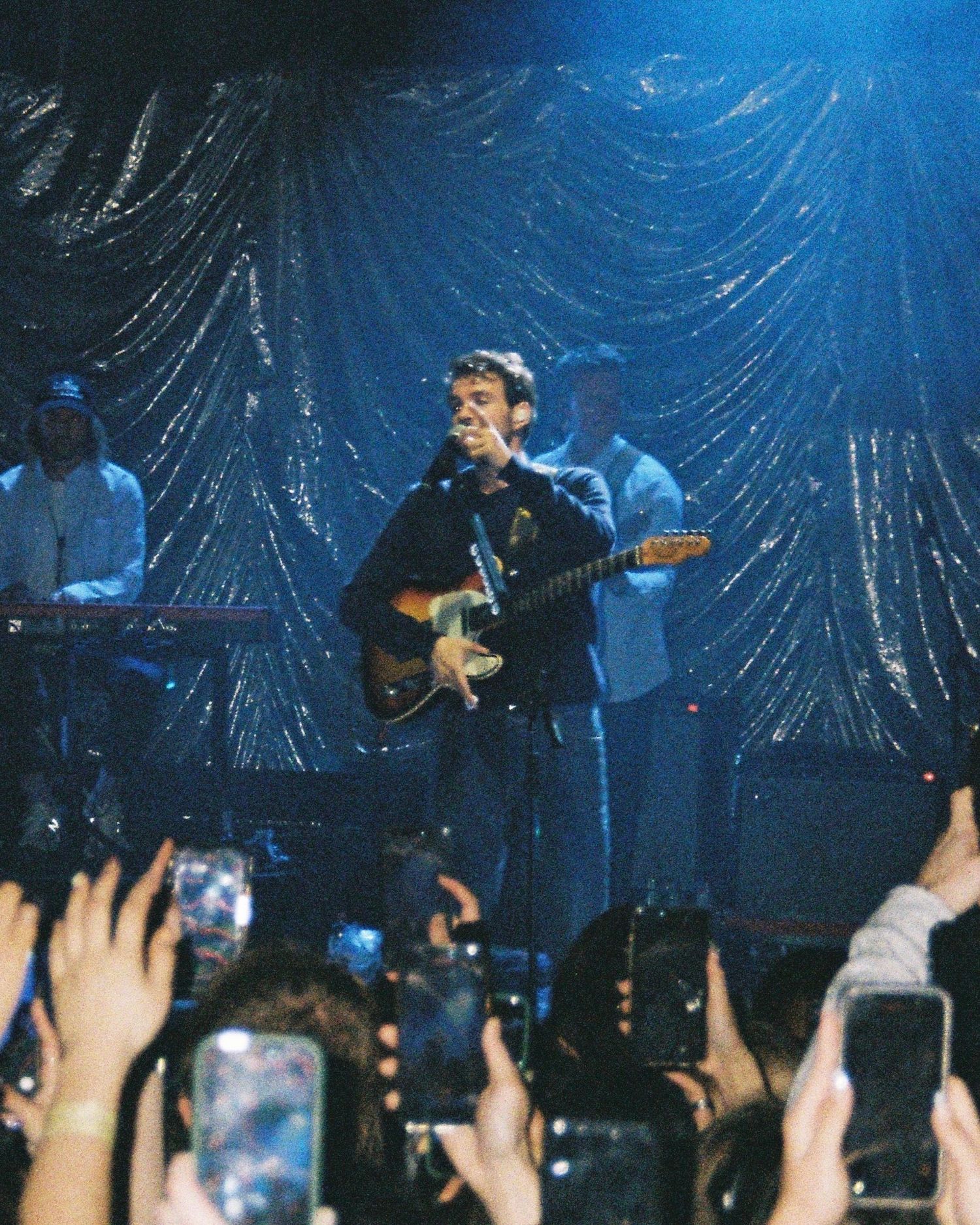 Musician playing guitar on stage with silver backdrop while crowd holds up phones recording.