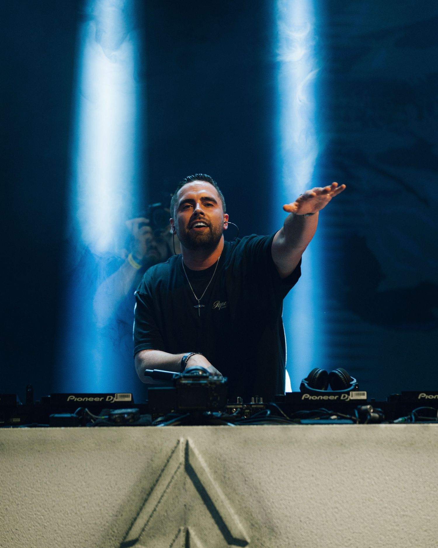 DJ performs behind decks with dramatic blue spotlight beams on dark stage.