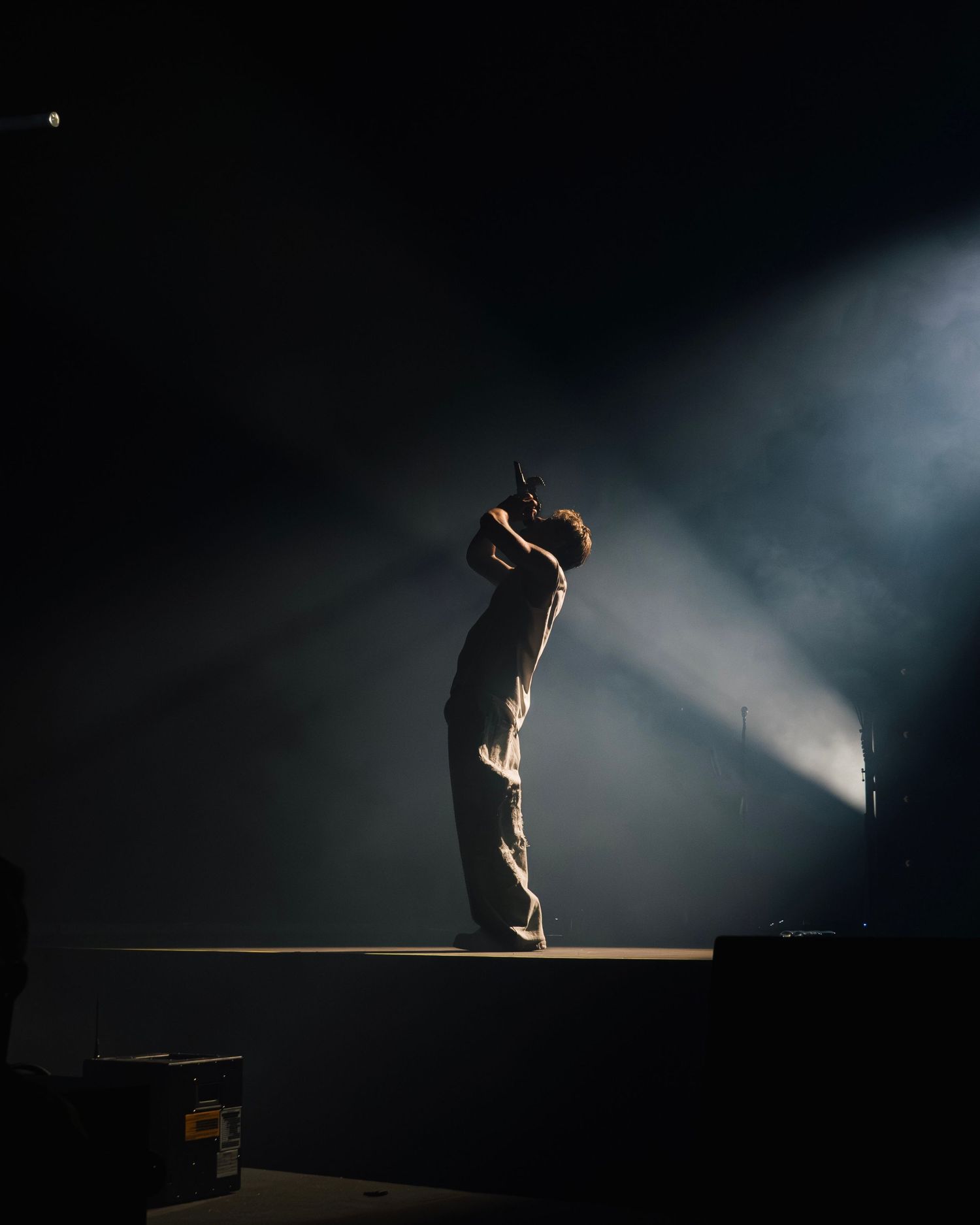 A performer stands alone on stage in dramatic silhouette, illuminated by striking beams of light cutting through smoke.