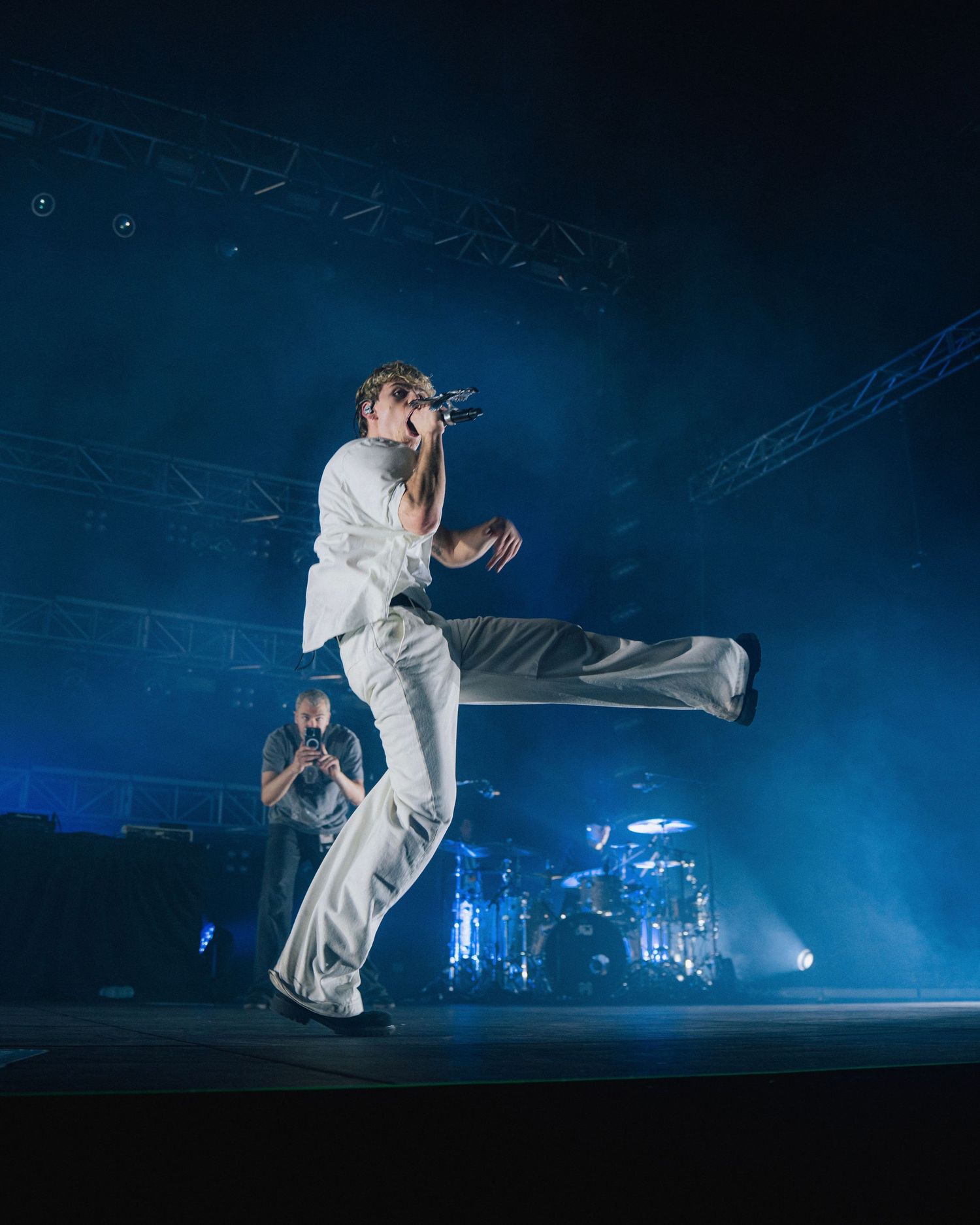 Performer in all-white outfit leaps energetically on stage with dramatic blue lighting and fog effects in concert setting.