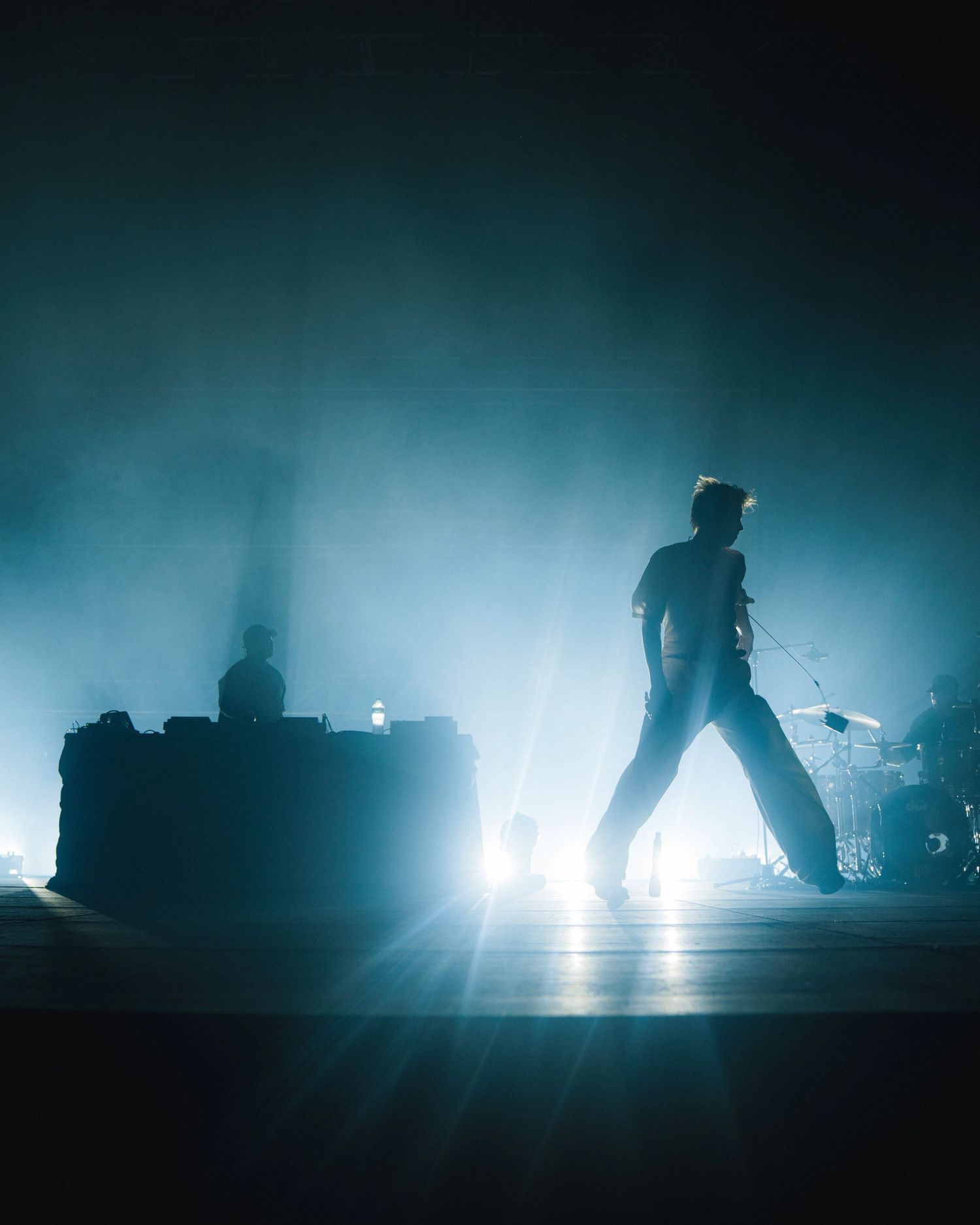 Silhouetted performer stands on stage in dramatic blue lighting with fog effects creating an atmospheric concert scene.