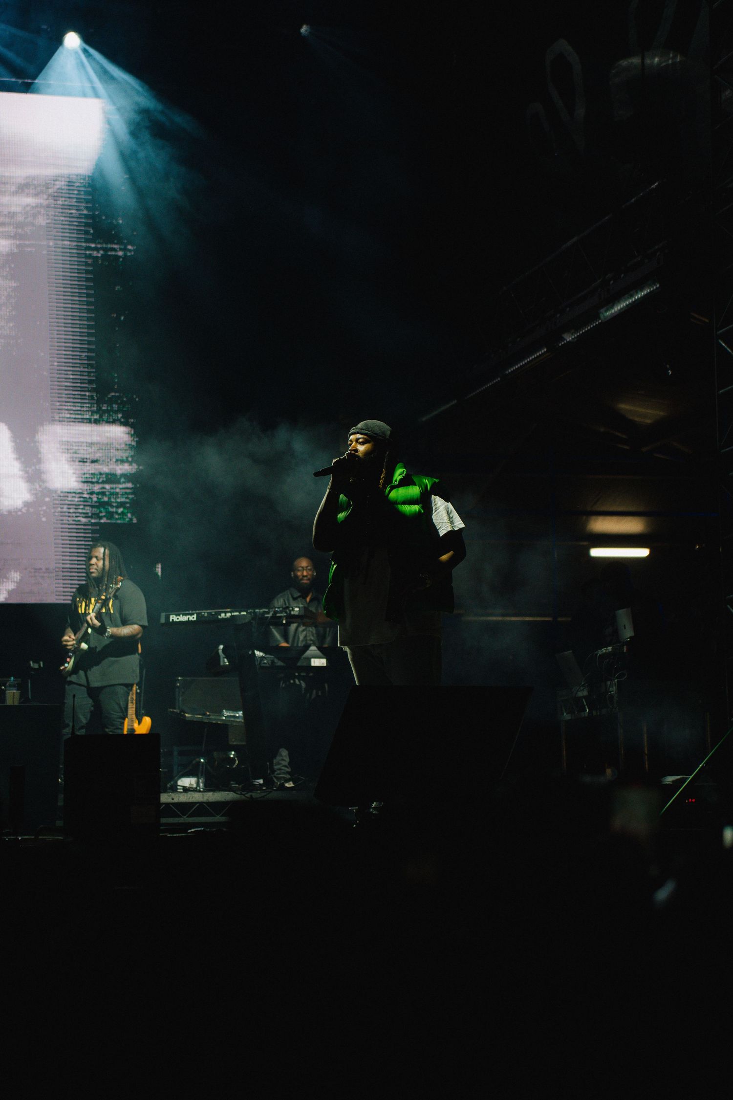 Dark concert stage with performer in neon green accent illuminated by dramatic spotlight through atmospheric haze.