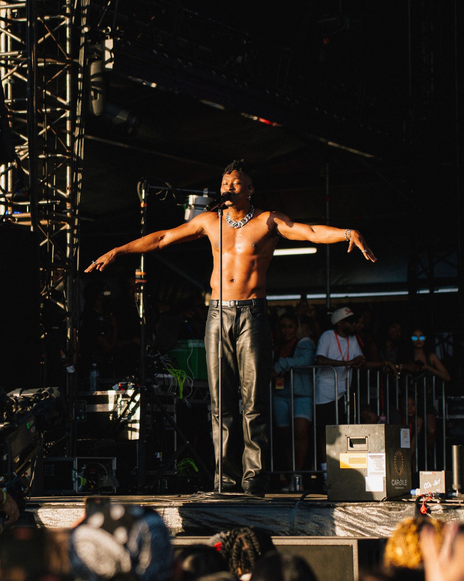 A performer stands shirtless with arms outstretched on stage during a concert in dramatic lighting.
