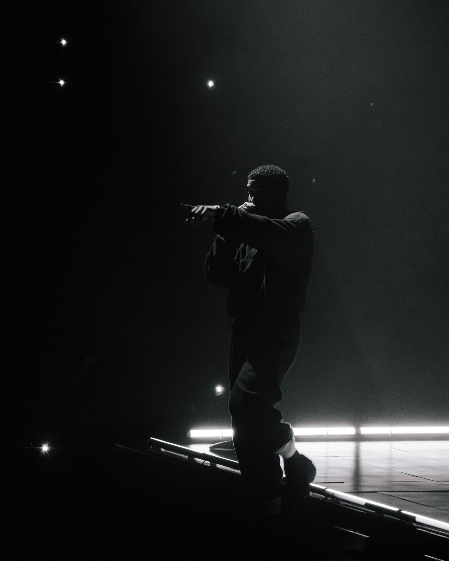 A silhouetted performer stands on stage in a dramatic black and white shot with spotlights creating a moody atmosphere.