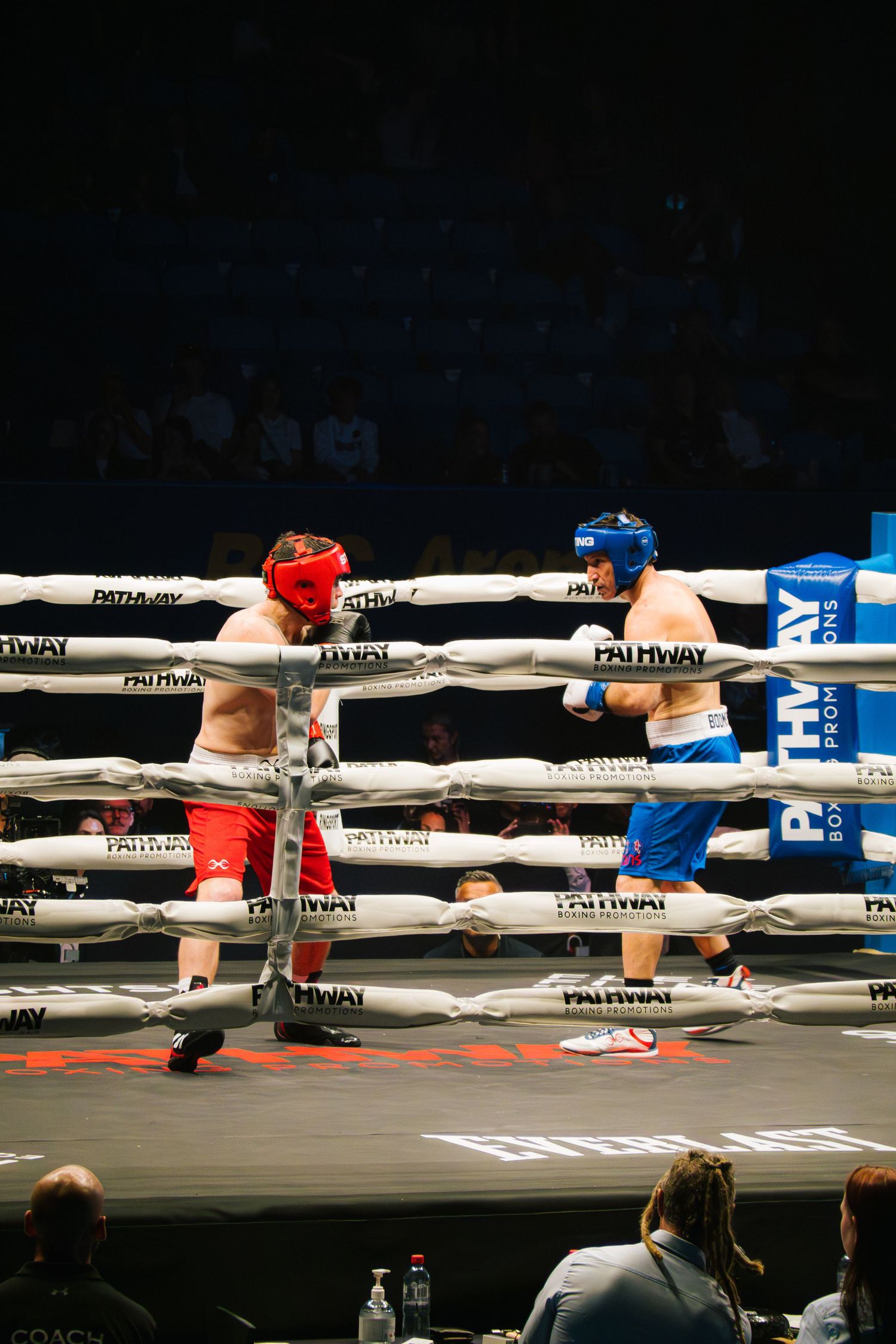 Two boxers in protective headgear compete through the ropes of a boxing ring.