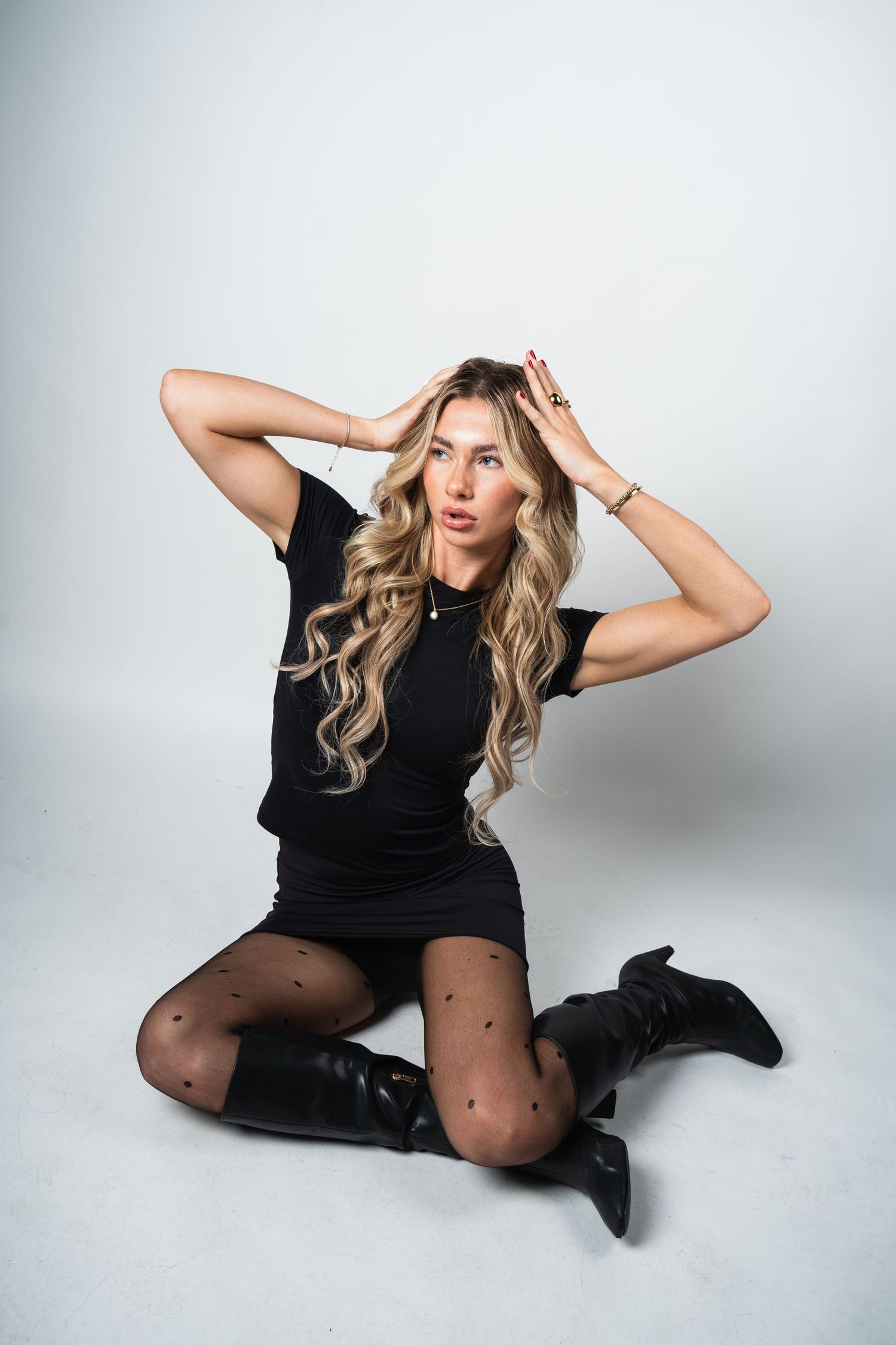 Fashionable model in black dress, patterned tights and boots poses against white studio backdrop.