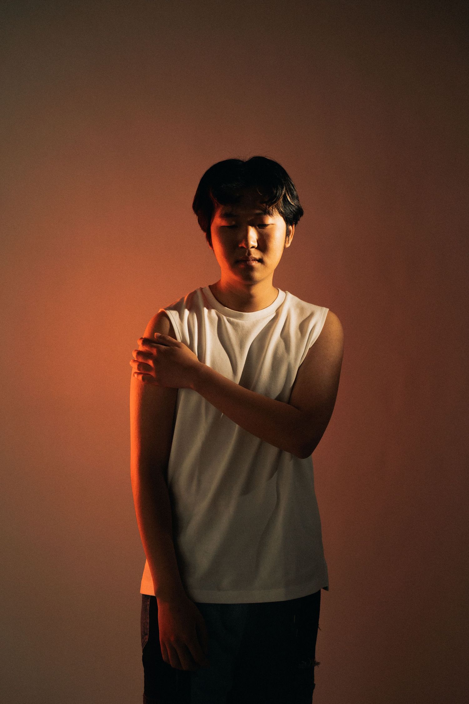 Portrait of person in white sleeveless shirt standing against warm orange-tinted background with soft lighting.