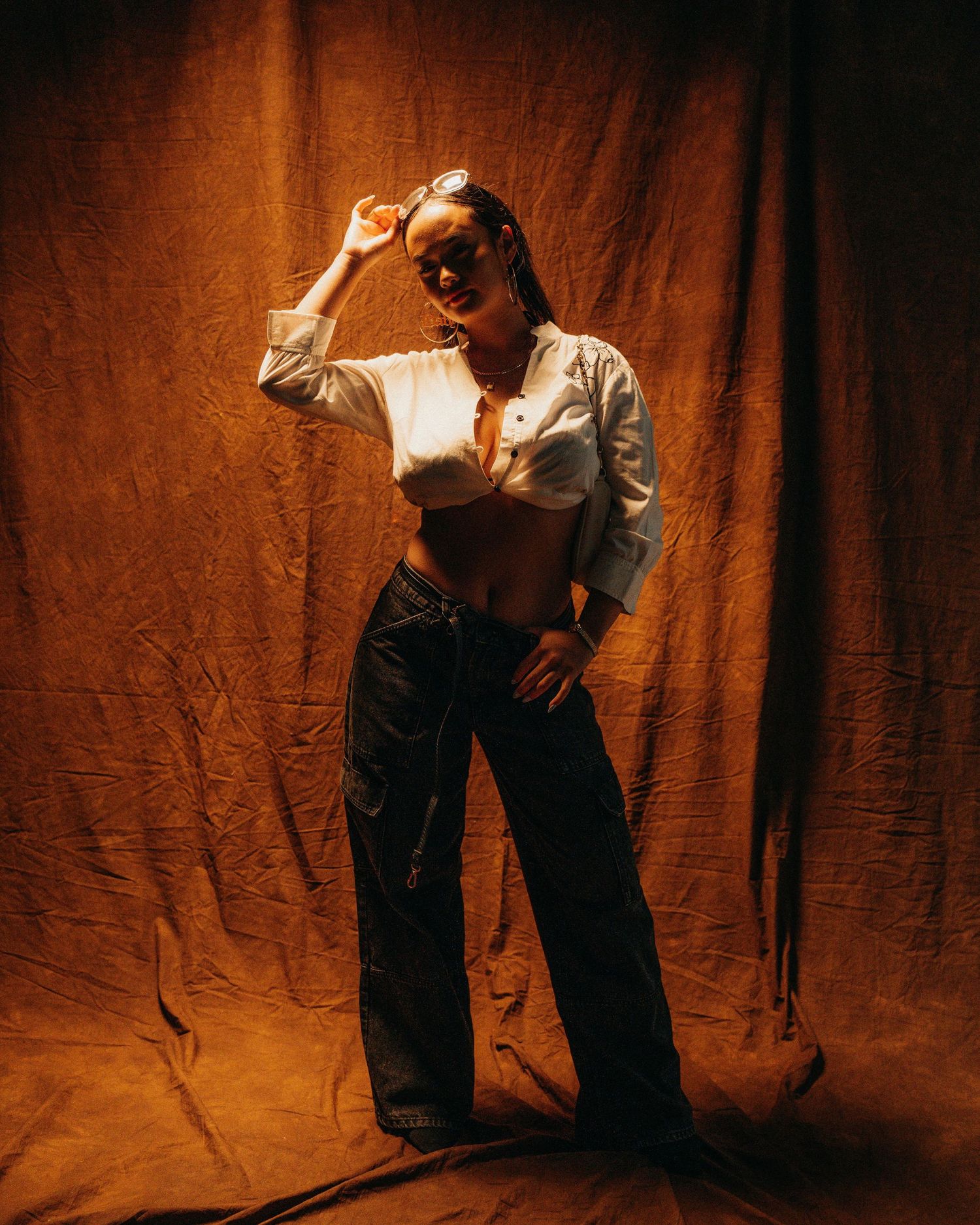Fashion portrait of person in white crop top and dark pants against brown backdrop.
