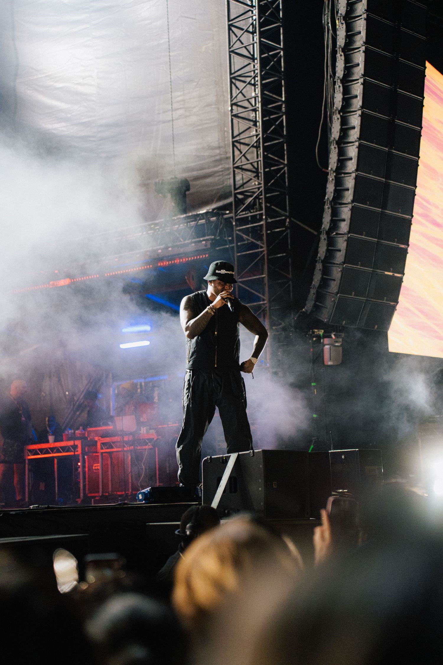 A performer on stage silhouetted in dramatic fog and lighting during a concert performance with large speaker arrays visible.