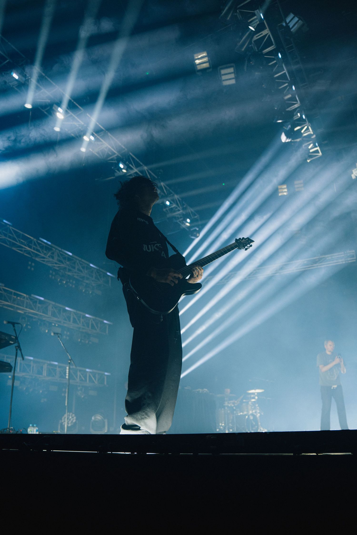 Silhouetted guitarist performs on stage with dramatic blue lighting and beaming spotlights creating atmospheric effect.