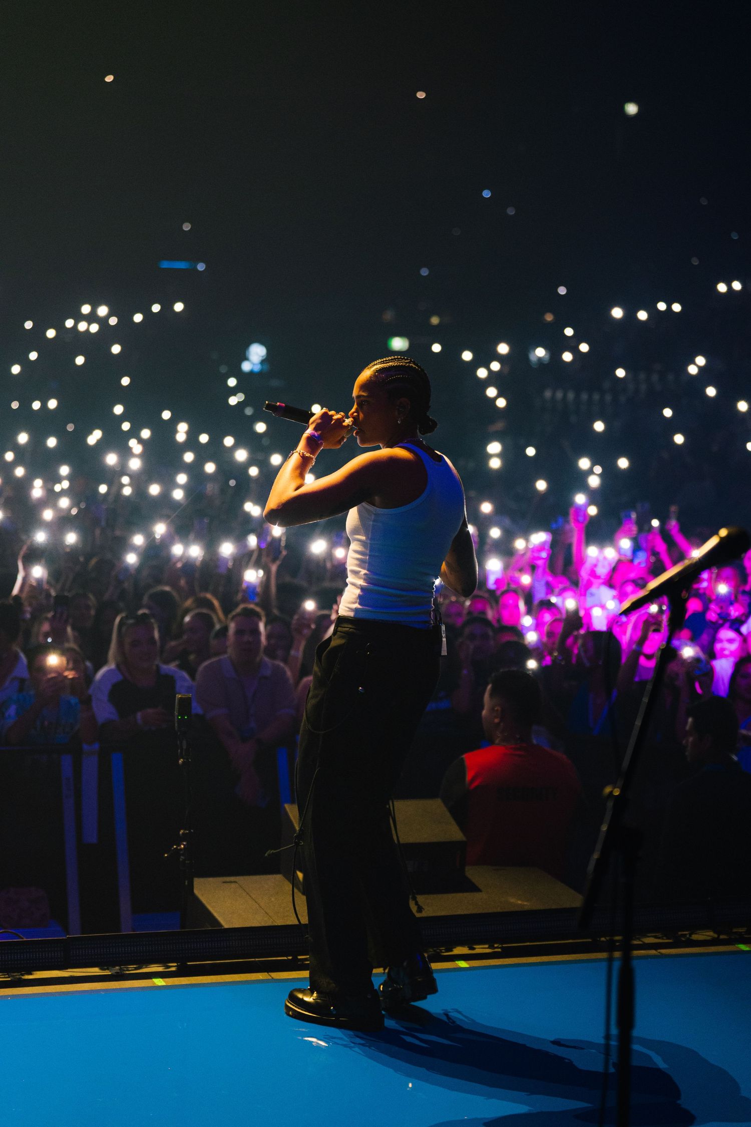 A performer in a blue top stands on stage at a concert with glowing phone lights from the crowd creating a starry atmosphere.