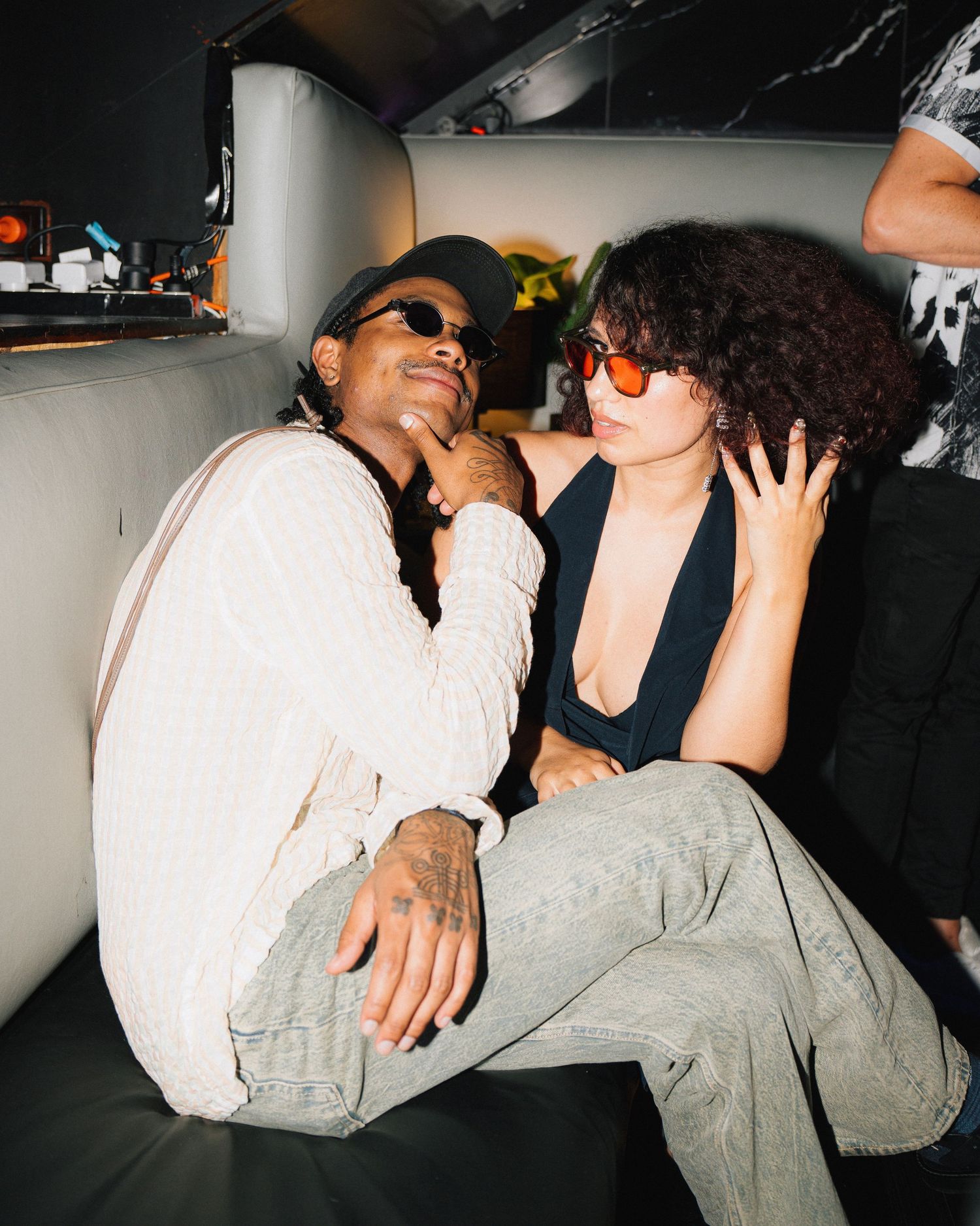 Two friends laughing and posing casually in a dimly lit setting wearing sunglasses and casual attire.