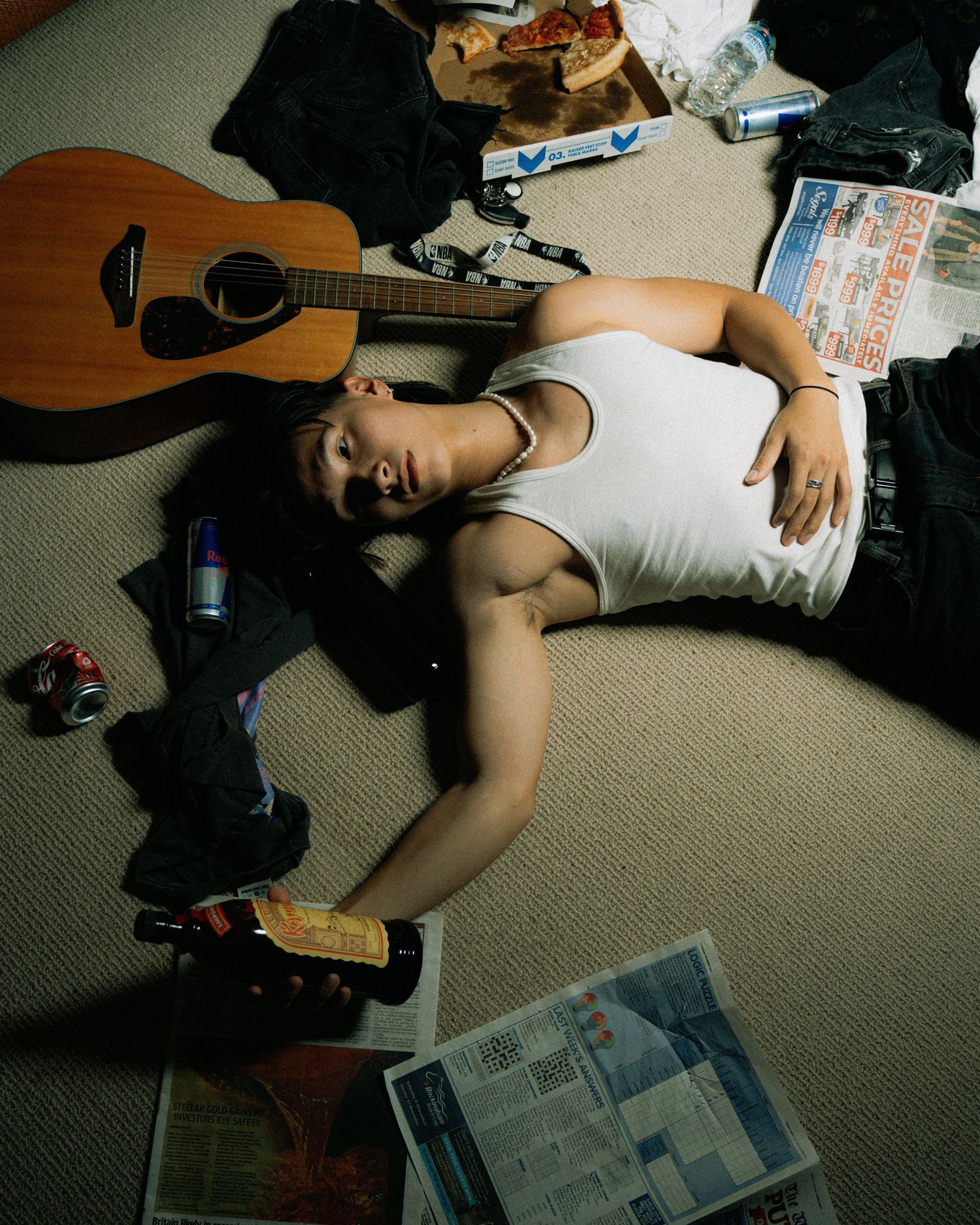 Someone lying on carpet surrounded by scattered newspapers and an acoustic guitar.