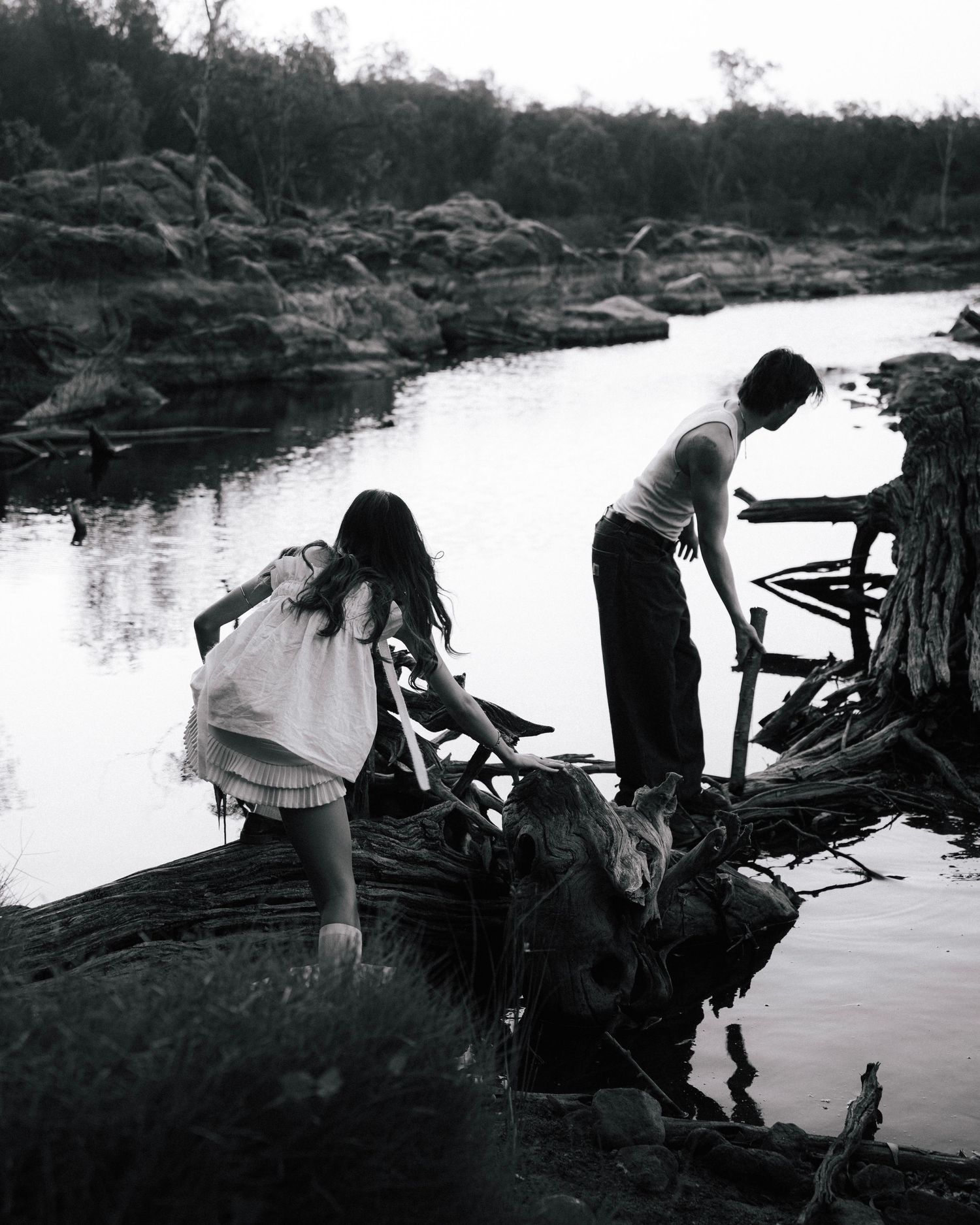 Two people explore a rocky waterfront and bend down to examine something by the water's edge in a black and white photograph.
