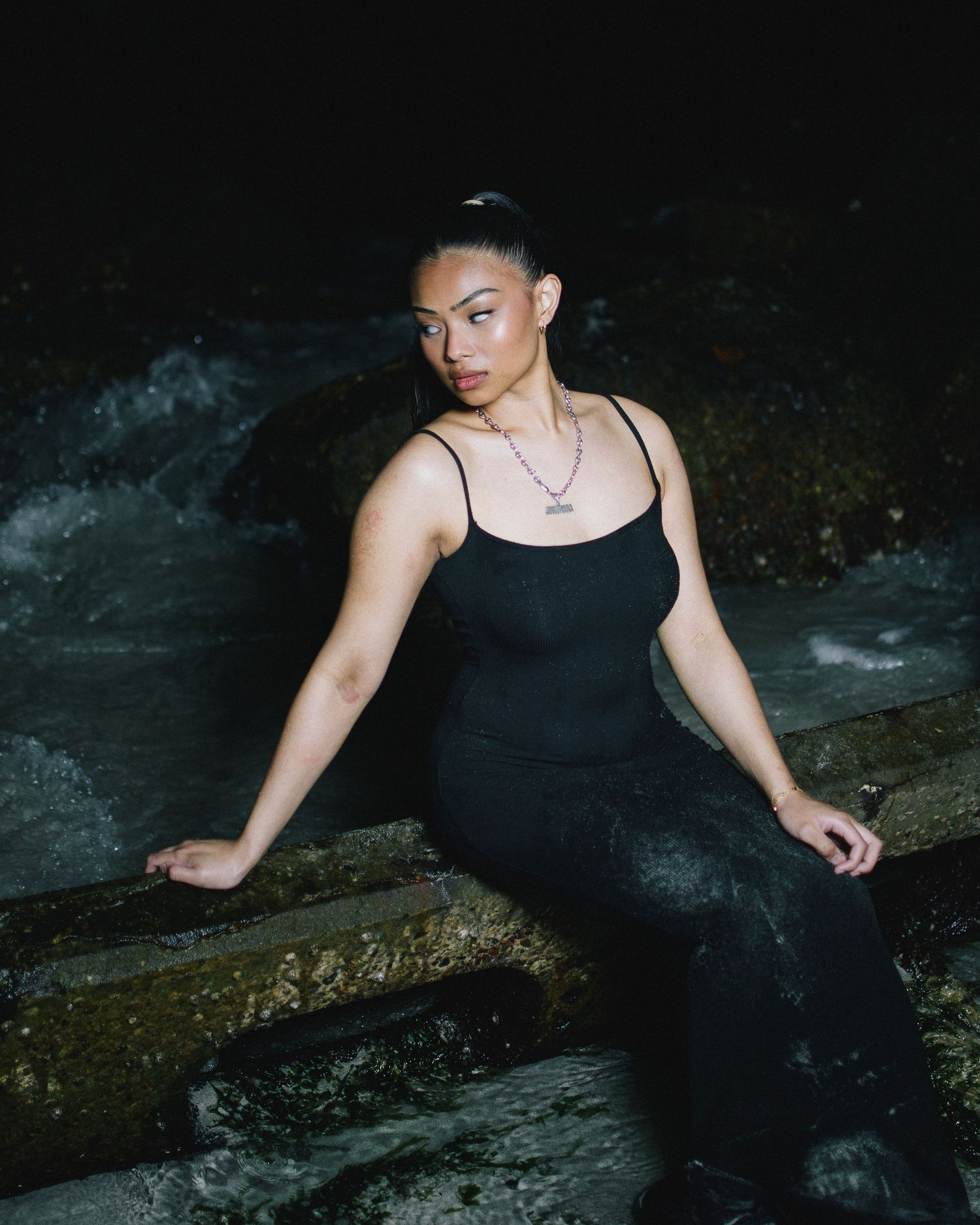A person in a black dress sits on rocky terrain near flowing water at night, with a dramatic dark background.