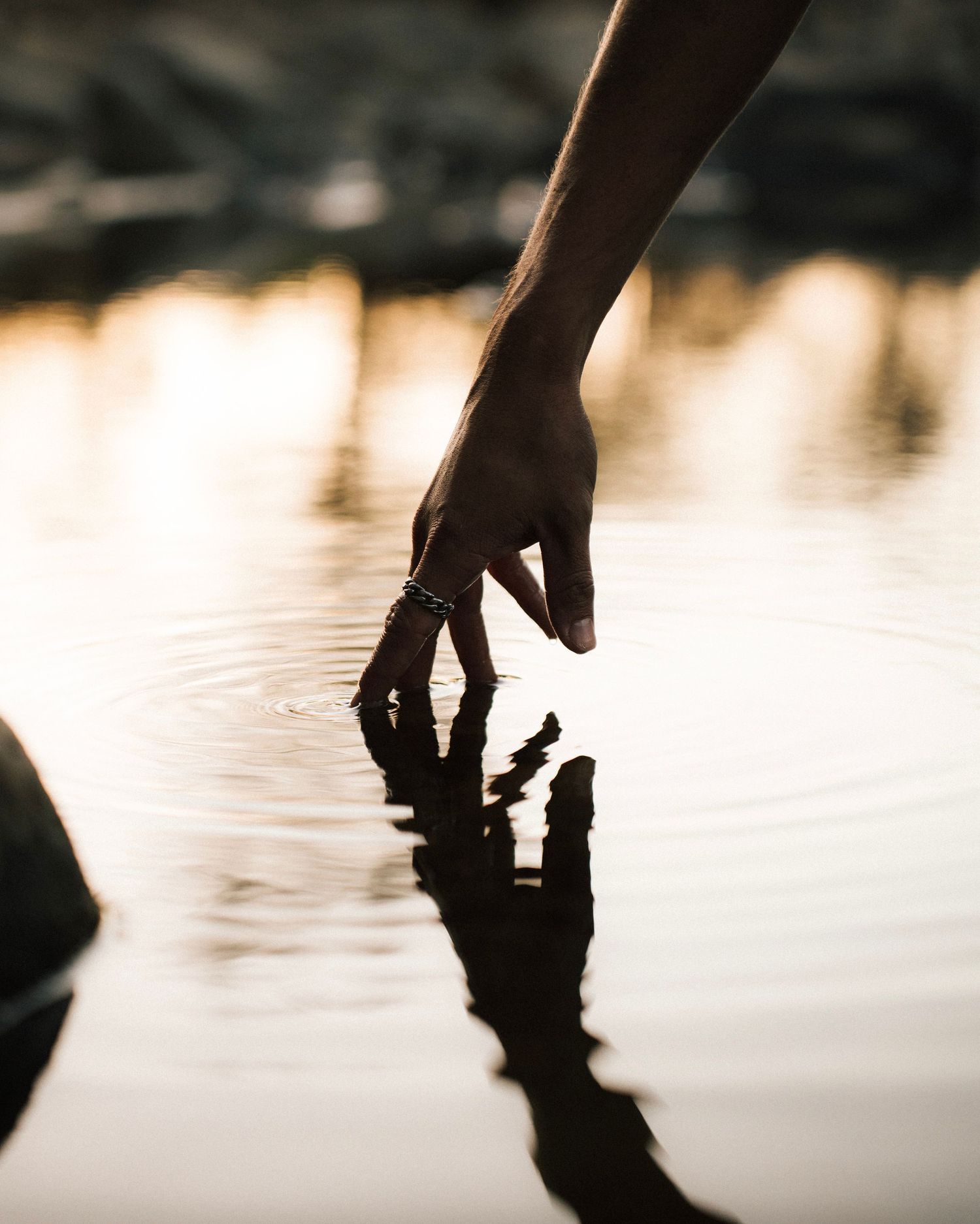 A silhouetted hand reaches down to touch the calm water surface creating a mirror-like reflection at sunset.