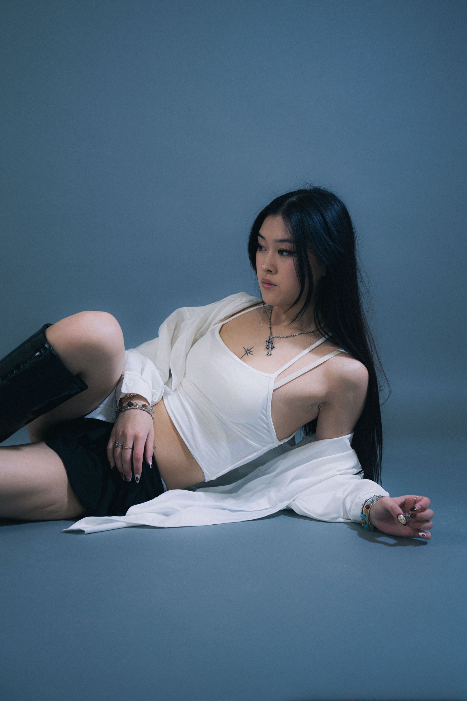Person in white crop top and sheer outer layer poses dramatically on gray studio floor against blue background.