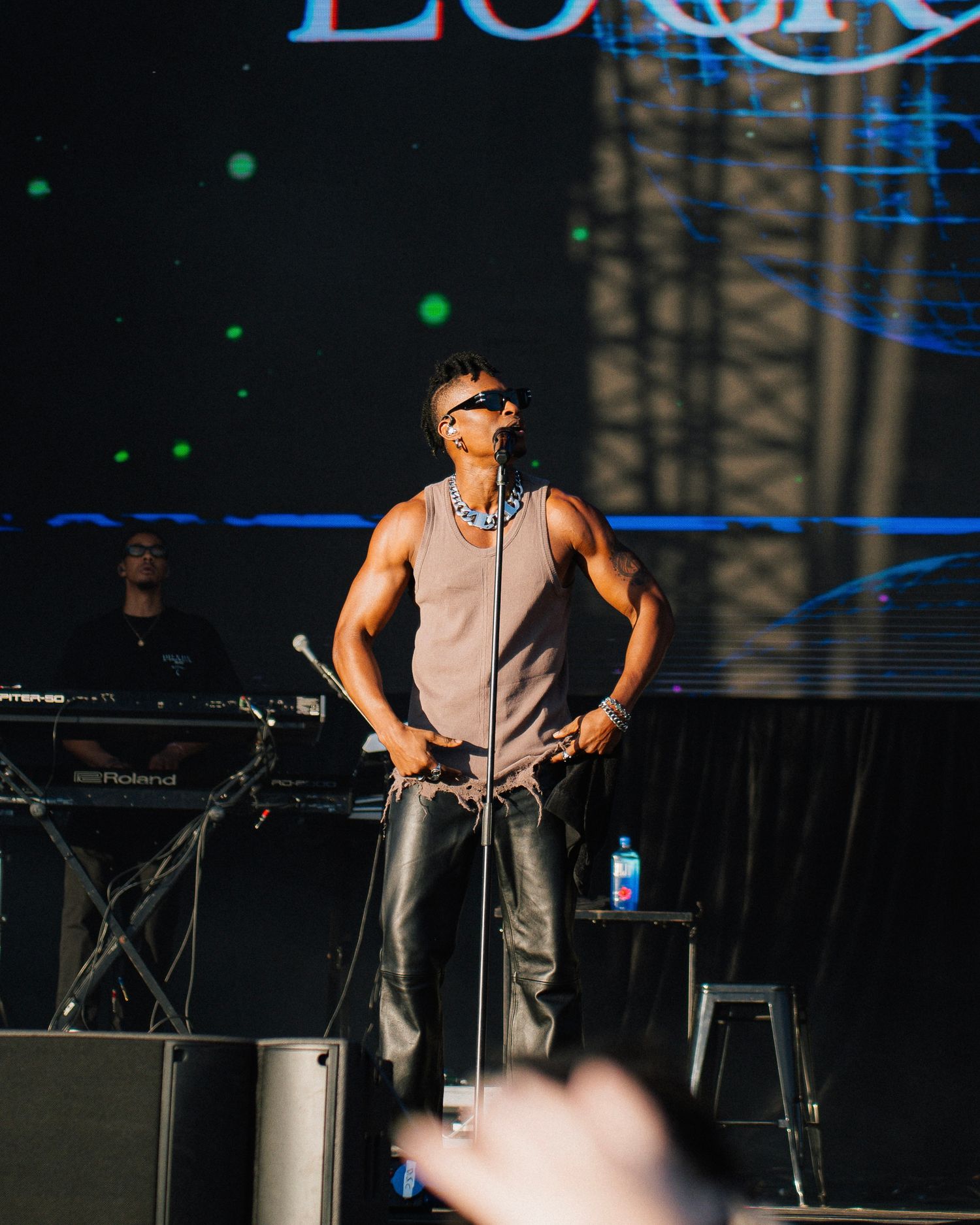 A performer in a gray tank top and sunglasses stands at center stage during an outdoor concert performance.