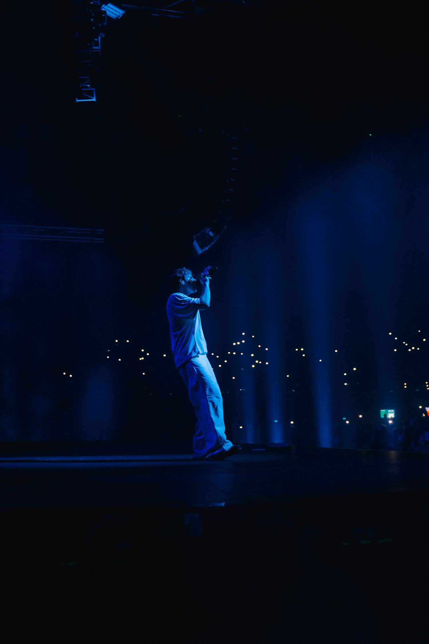 Performer silhouetted in blue stage lighting stands against backdrop of distant city lights during nighttime show.