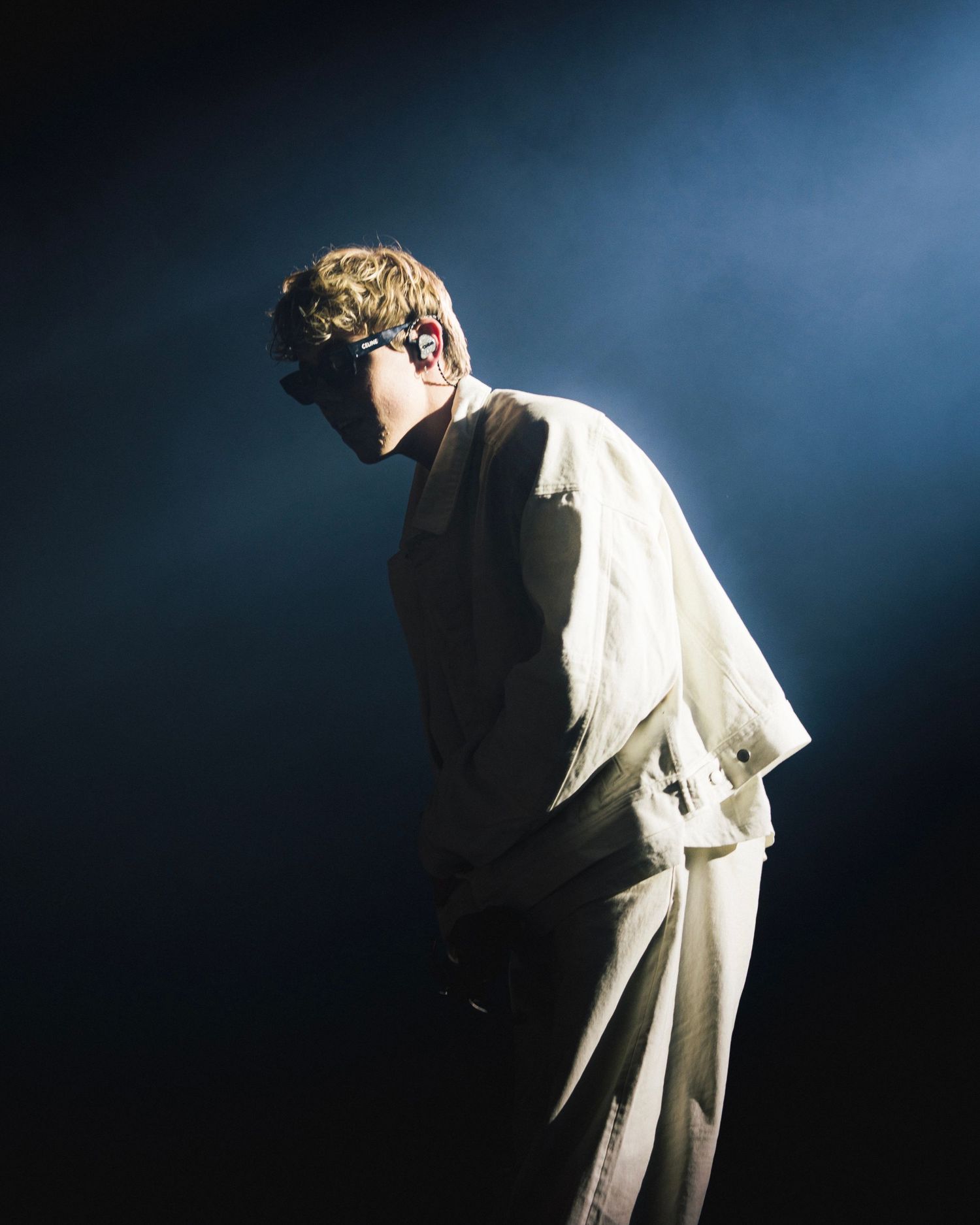 Figure in white clothing stands in dramatic lighting against dark background during performance on stage.