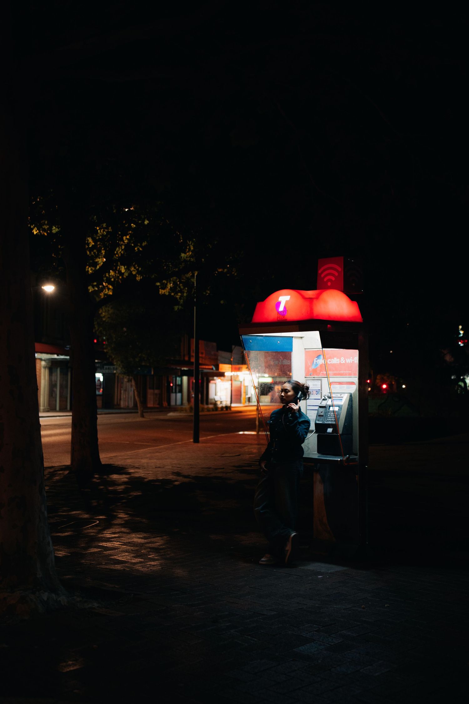A red-topped illuminated bus shelter glows on a dark street at night, with a silhouette of a person waiting.