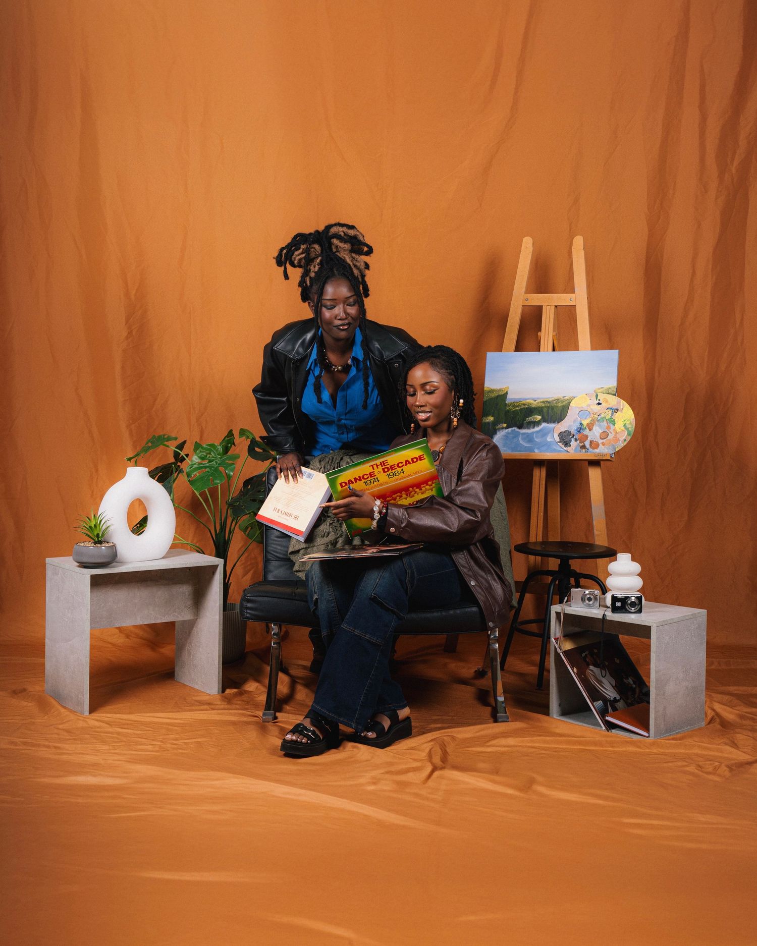 Two people sit together surrounded by art supplies and props against an orange backdrop in a creative studio setup.