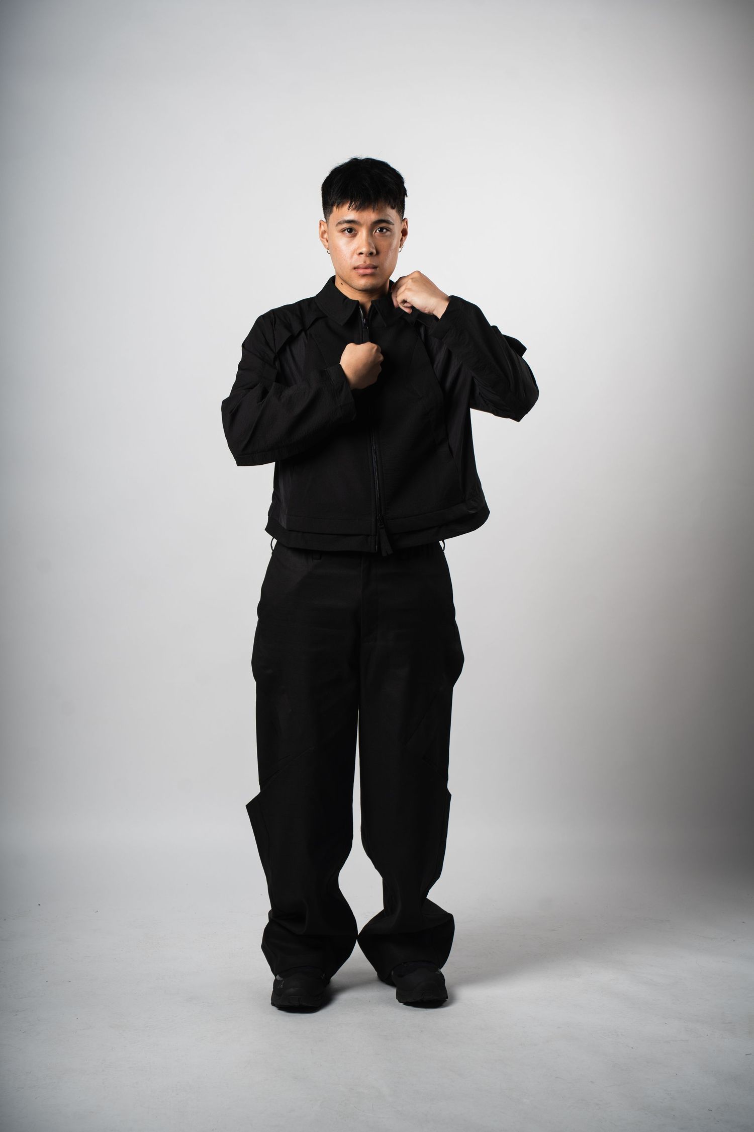 Person wearing all black martial arts uniform stands in fighting stance against light gray background.