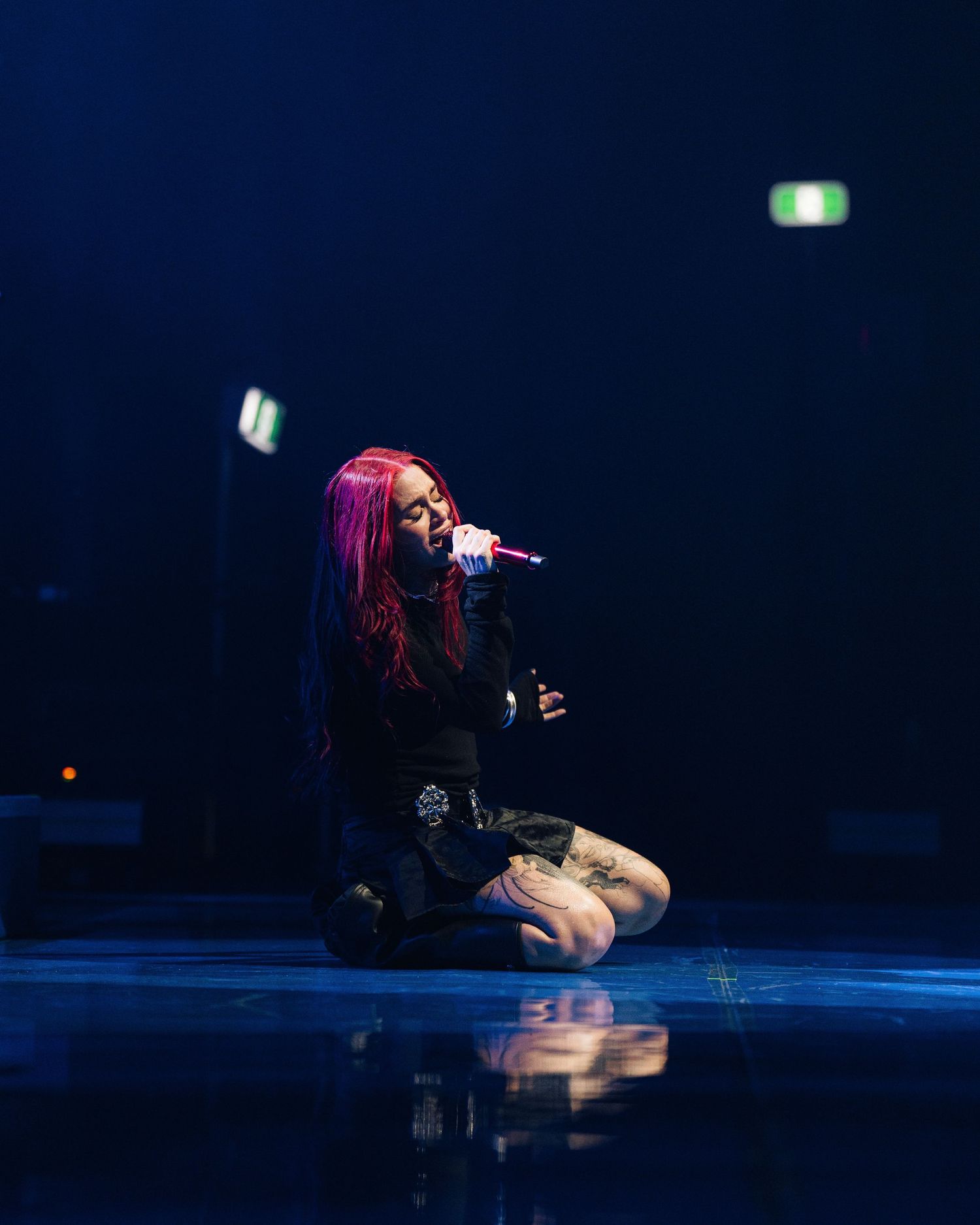 A performer with pink hair kneels on stage singing into a microphone during a concert with dramatic blue lighting.
