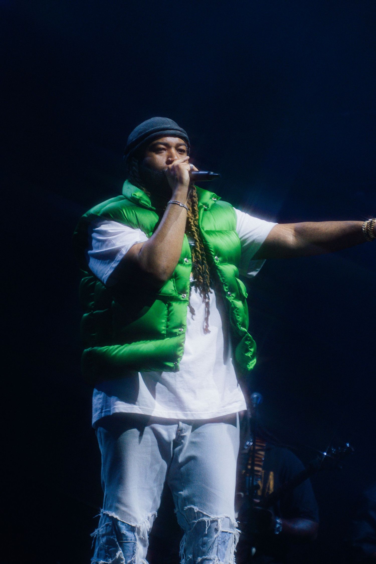 Performer wearing bright green vest, white shirt and ripped jeans holds microphone on dark stage with blue lighting.