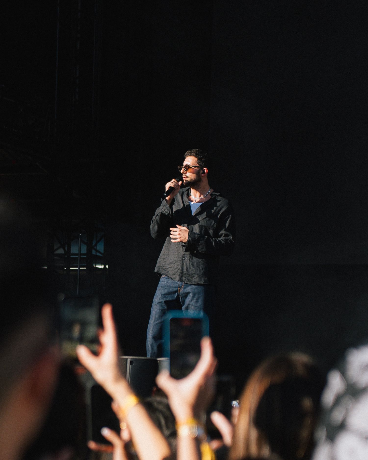 A performer in sunglasses and dark jacket stands on stage with microphone as crowd raises hands in silhouette.
