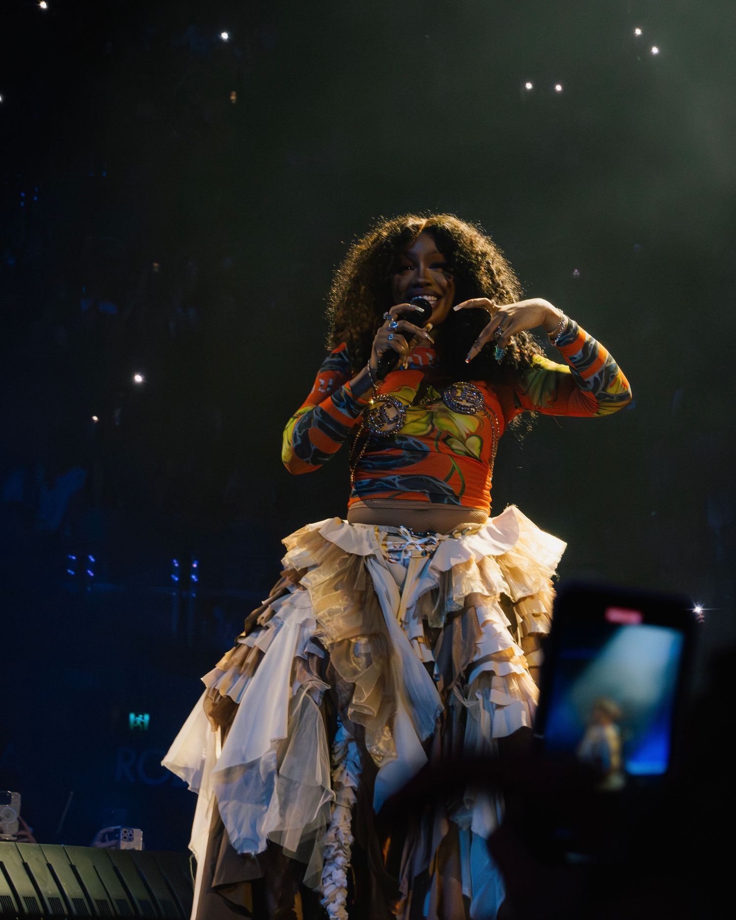A performer in an orange top and tattered white skirt costume dances energetically on stage under dramatic lighting.