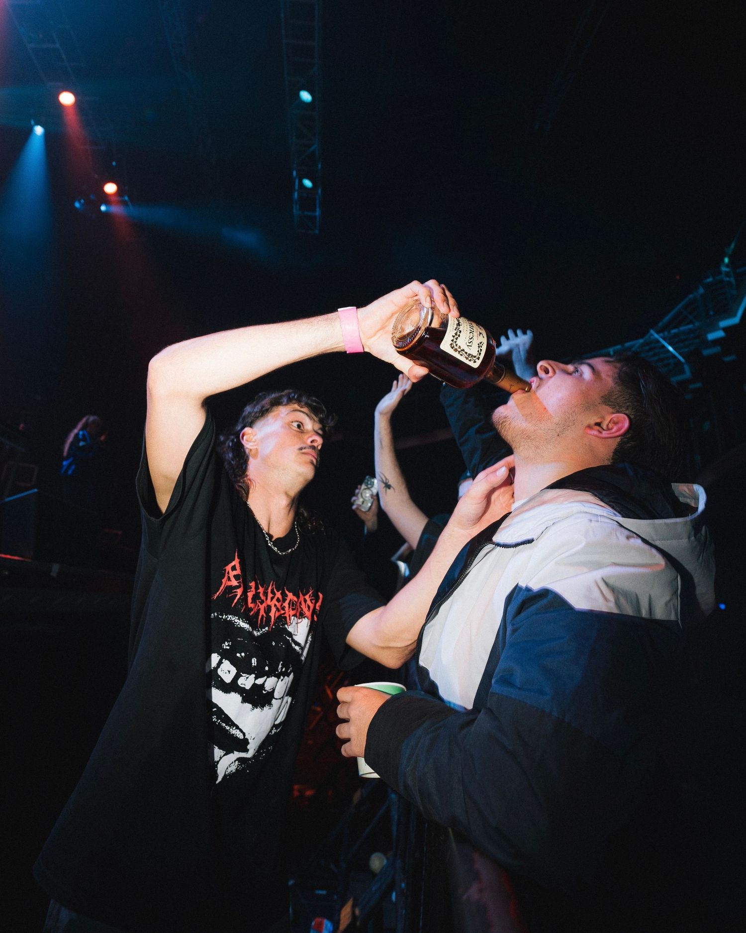 Two concert attendees sharing a drink in a darkened venue with dramatic lighting.