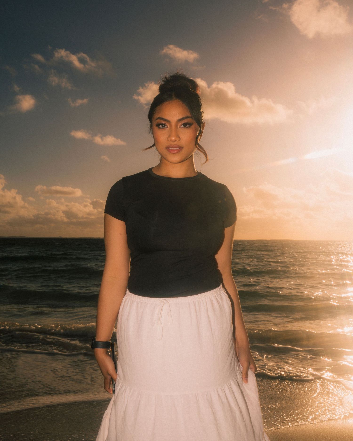 Figure in black top and white flowy skirt poses at sunset on a beach with golden clouds and ocean waves.