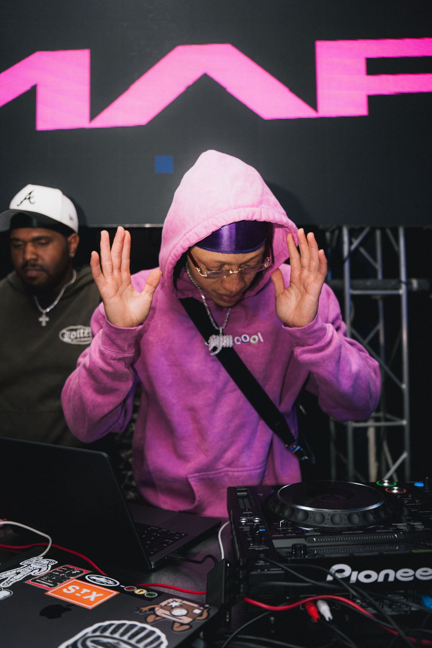 DJ in purple hoodie performing under pink MAF signage with Pioneer equipment.