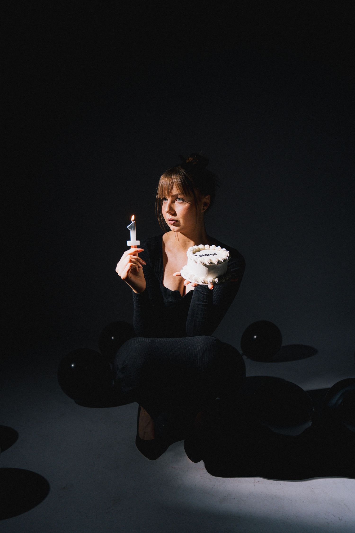 A person in dark clothing sits in moody lighting holding a candle against a dark background.