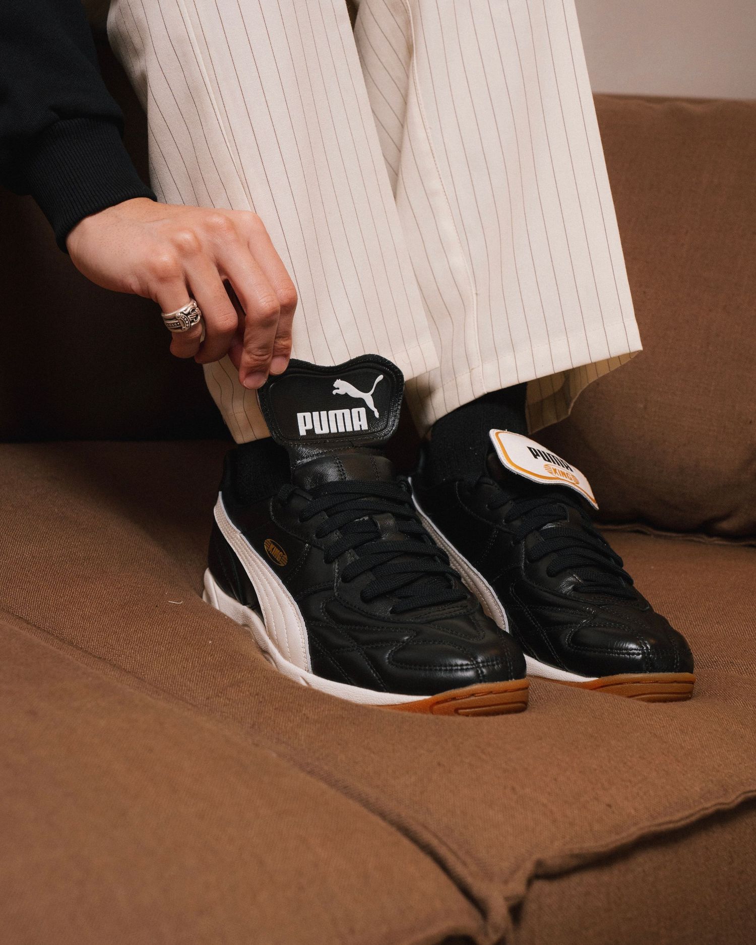 Person wearing striped pants sits on brown couch showing off black and white Puma sneakers with gum soles.