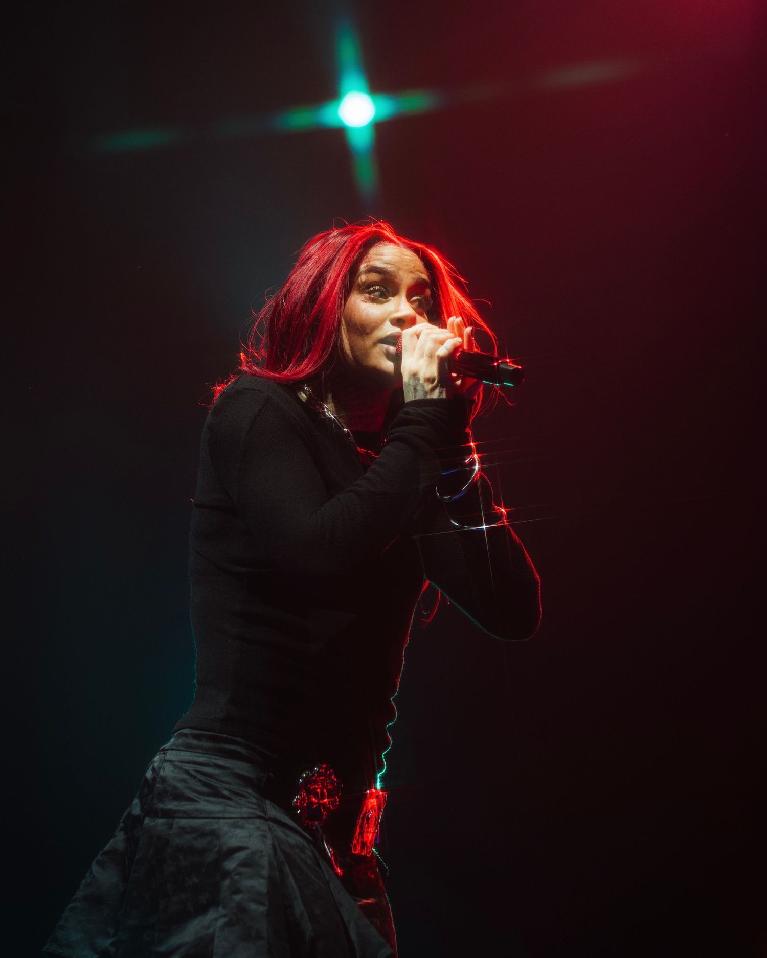 Singer with bright red hair performing on dark stage with dramatic lighting effects.