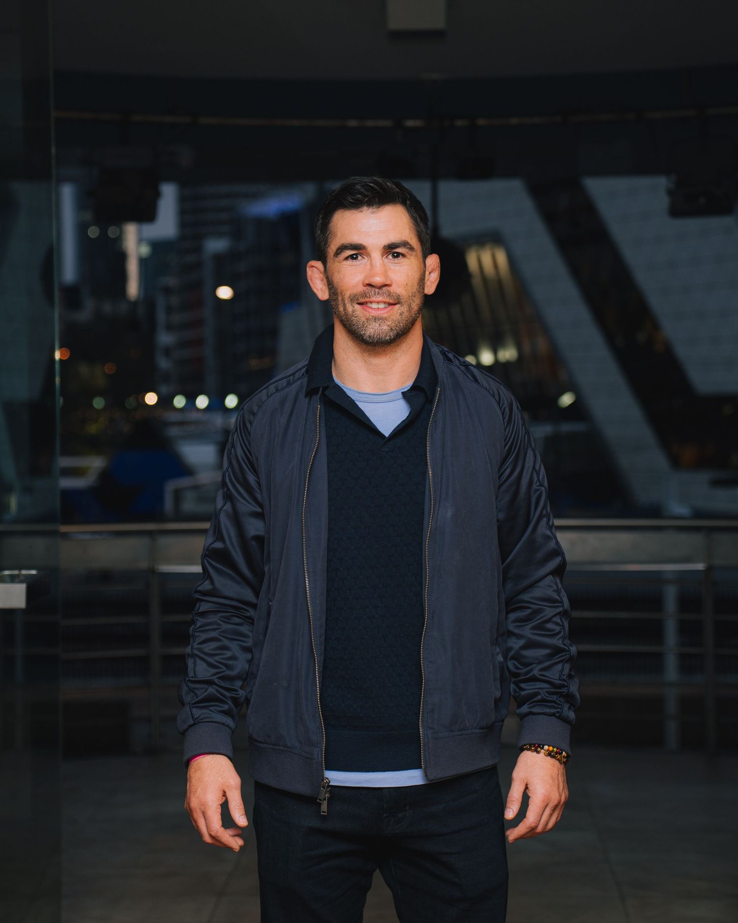 Man in casual navy jacket and sweater stands outdoors at night.