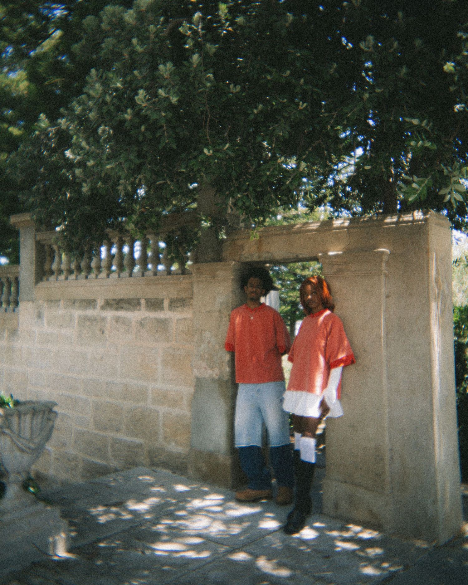 Two people standing against stone wall in casual outfits under dappled tree shadows.