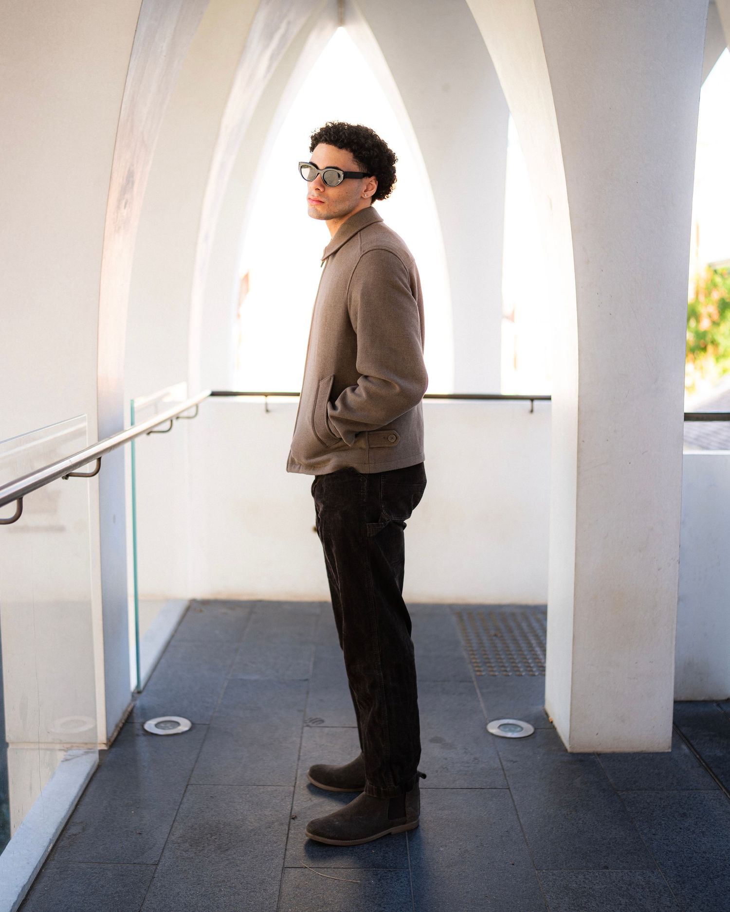 Stylishly dressed person in glasses standing in an arched white hallway wearing beige jacket and dark pants.