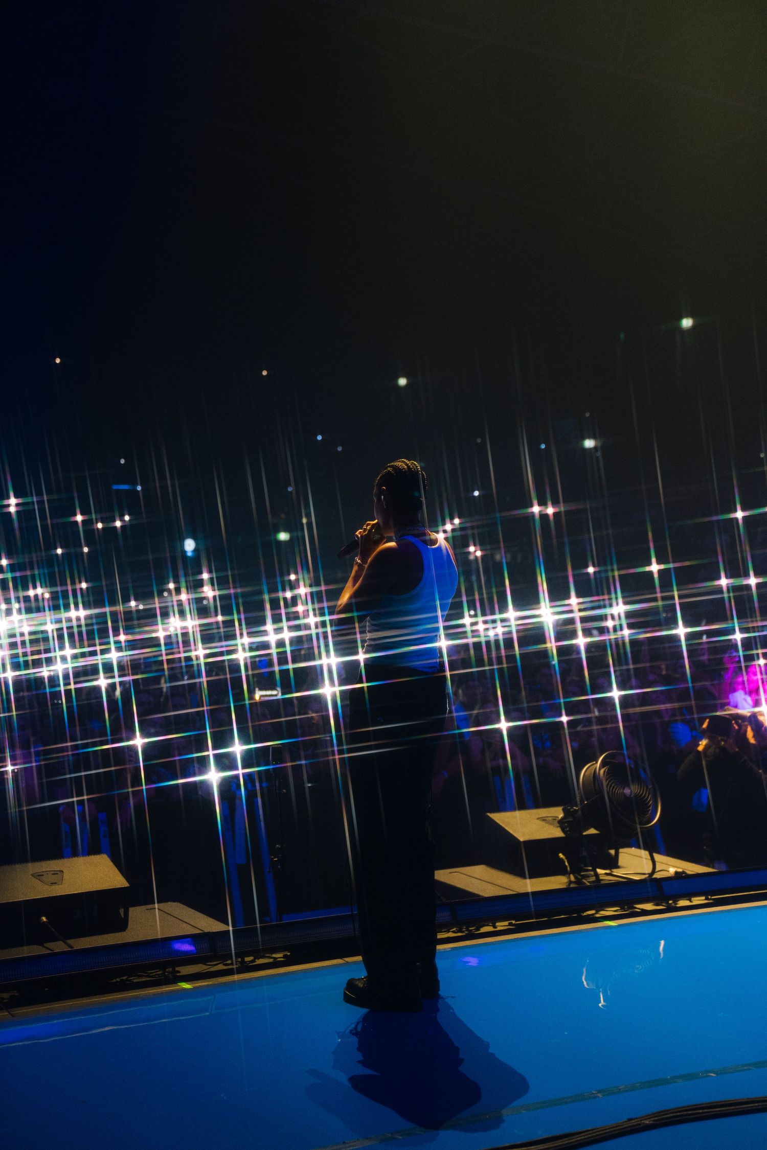 A silhouetted performer on a blue-lit stage against a backdrop of sparkling concert lights and reflections.