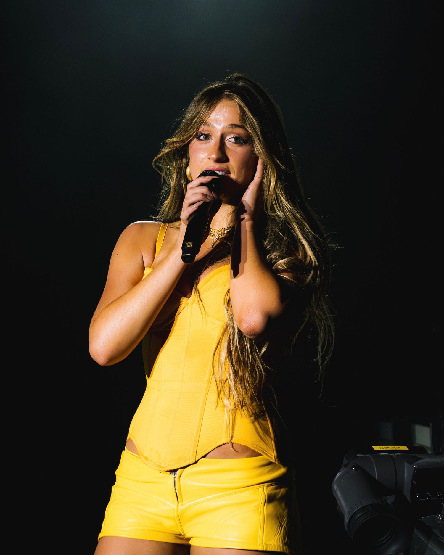 A performer in a bright yellow outfit sings passionately on stage against a dark background during a live concert performance.