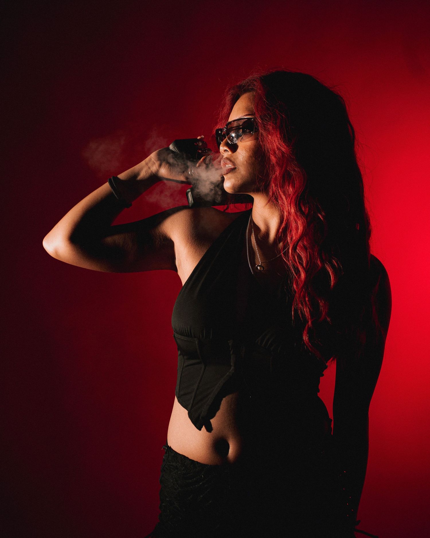 Person with red hair wearing black outfit drinks from bottle against dramatic red lighting background.