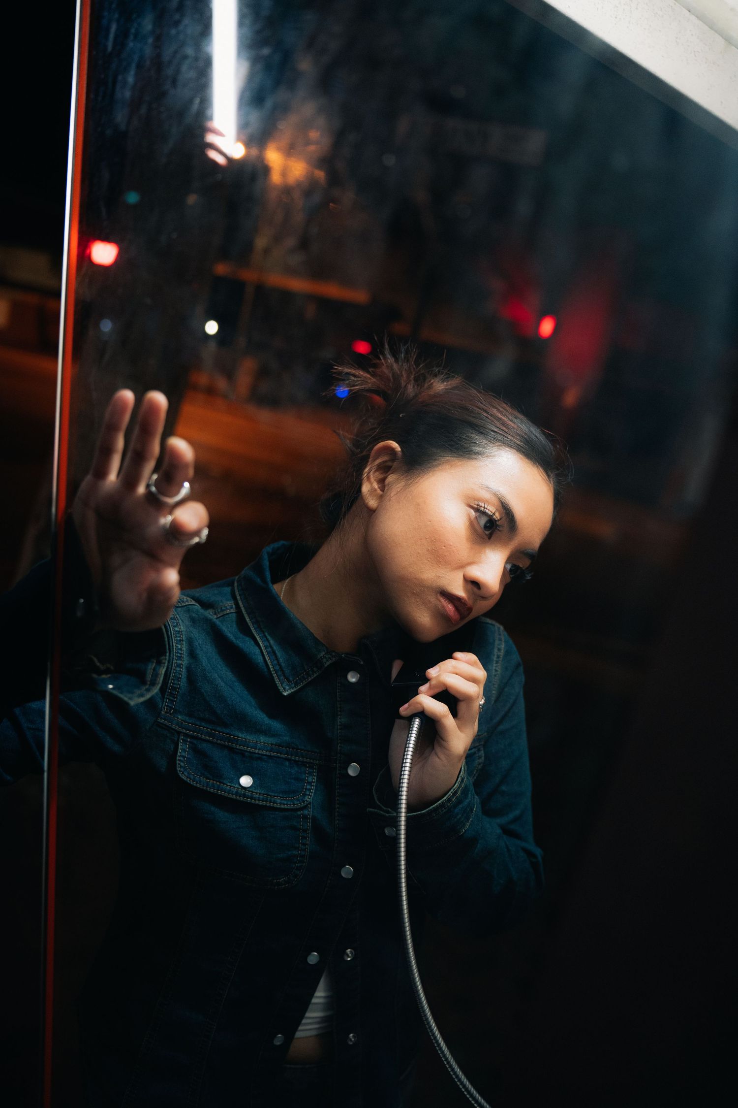 Person in denim jacket using vintage payphone booth at night with moody lighting and reflective glass.