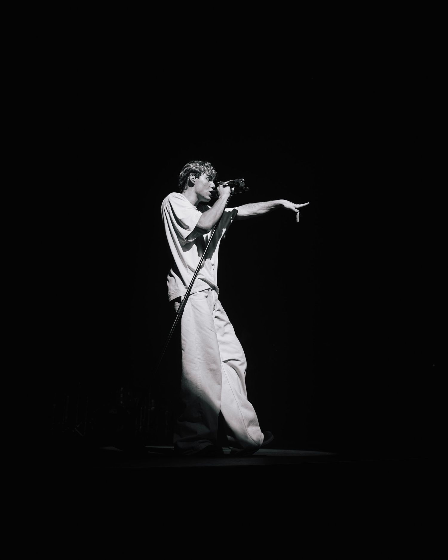 A performer on stage gestures while holding a microphone in a dramatic black and white spotlight shot.