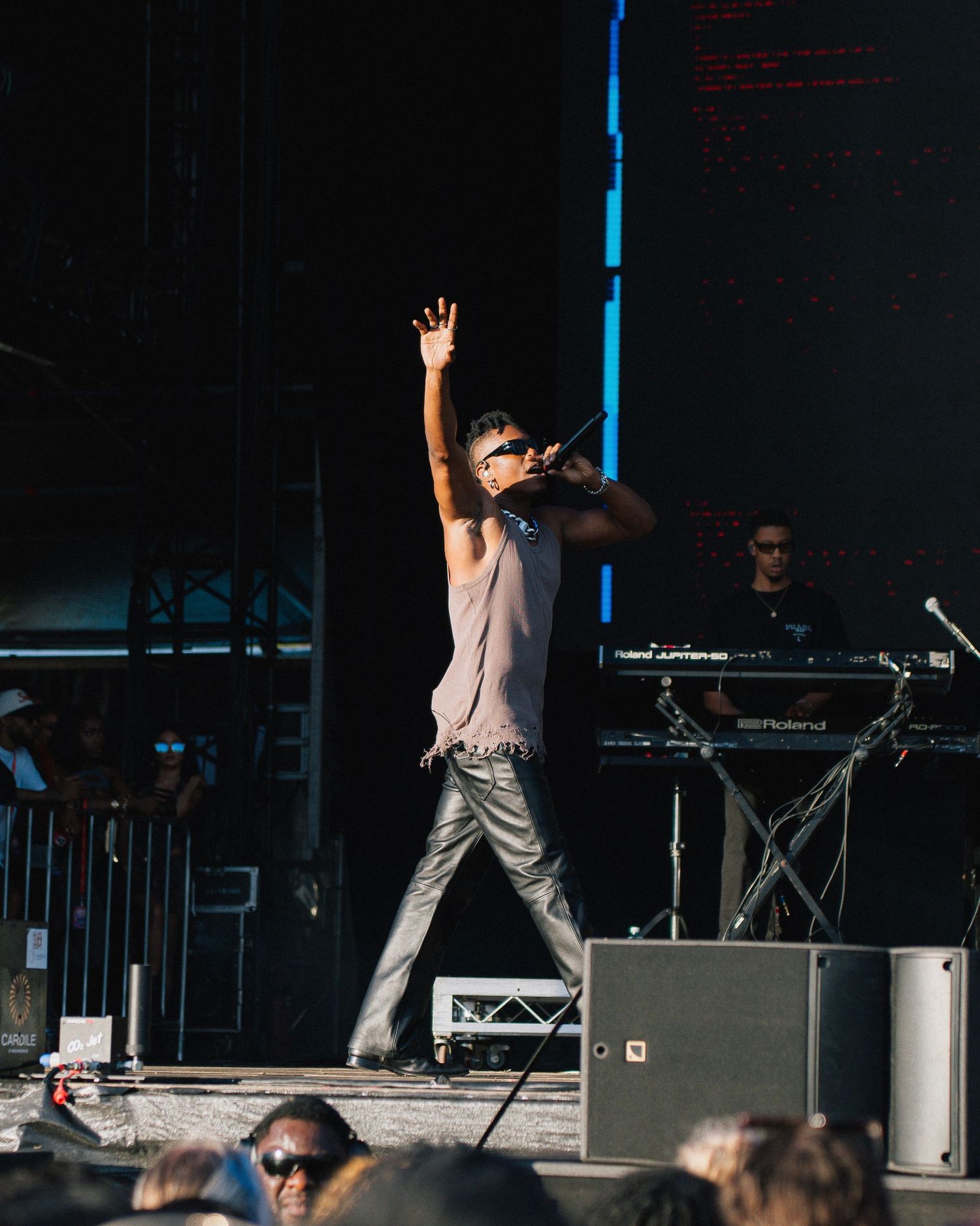 A performer stands energetically on stage during a concert, with keyboards and equipment visible in the background.