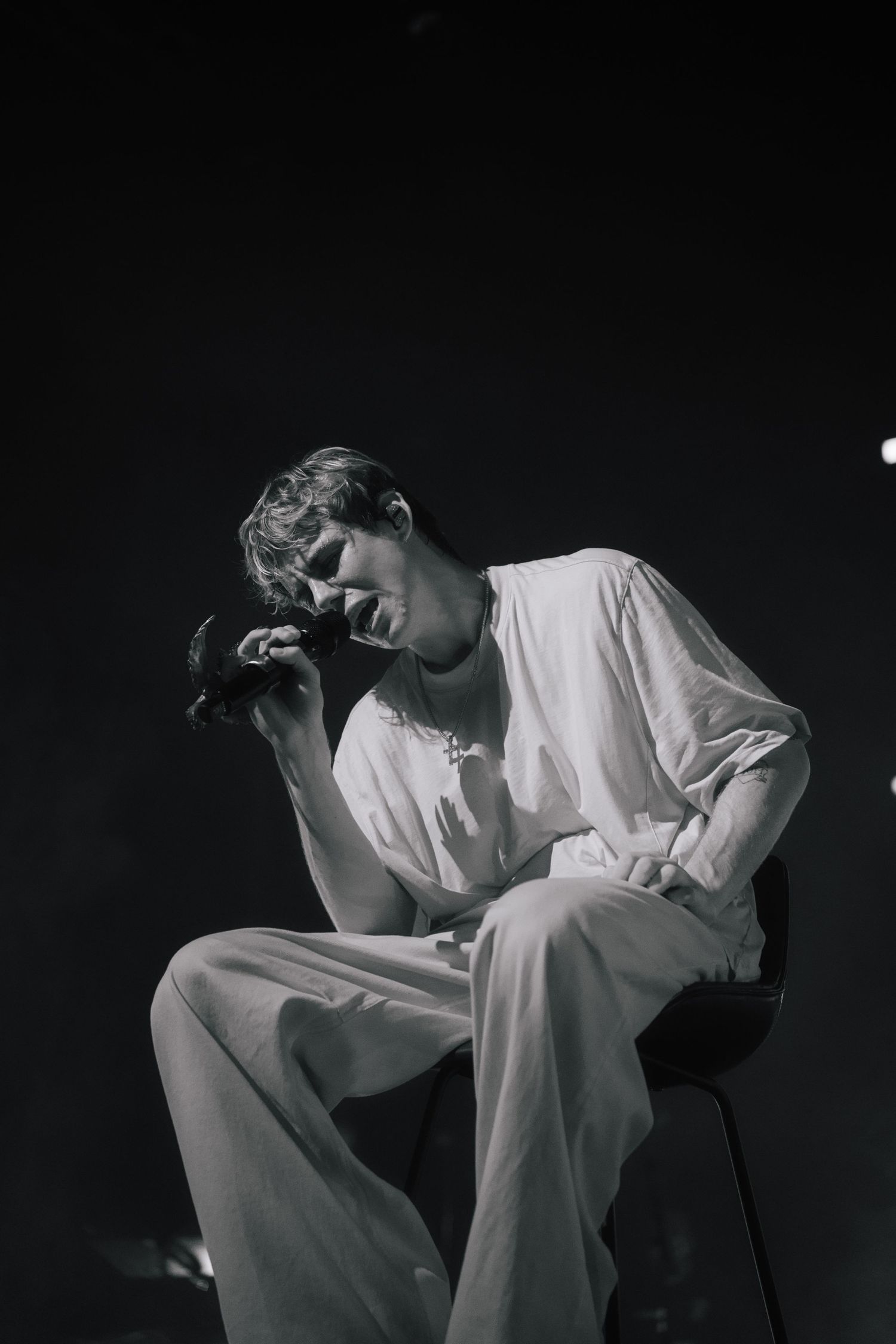 Black and white artistic portrait of a figure in all-white clothing sitting contemplatively on a chair against dark background.
