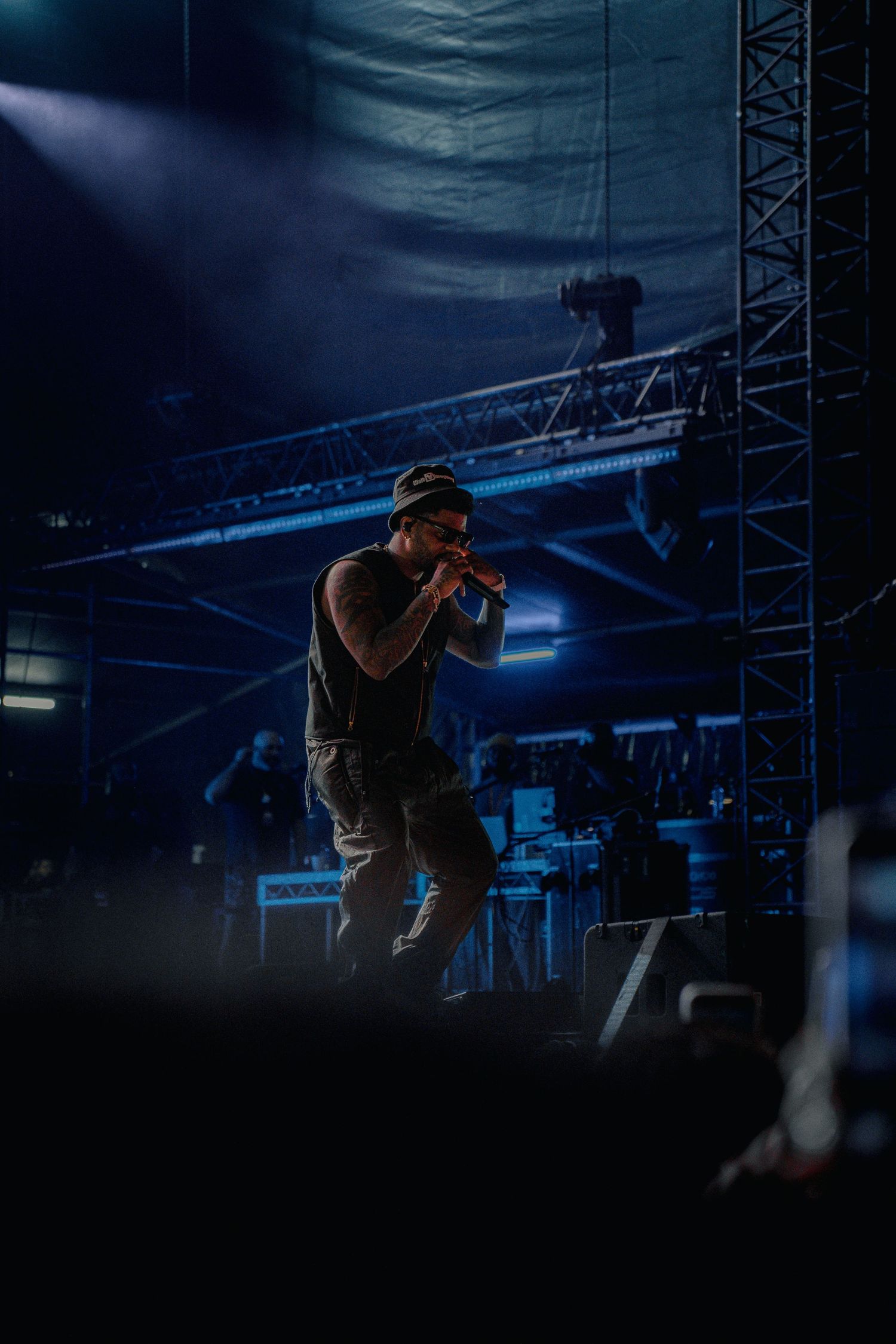 Silhouette of a performer in a baseball cap shown on dark stage with dramatic blue lighting and metal truss rigging above.