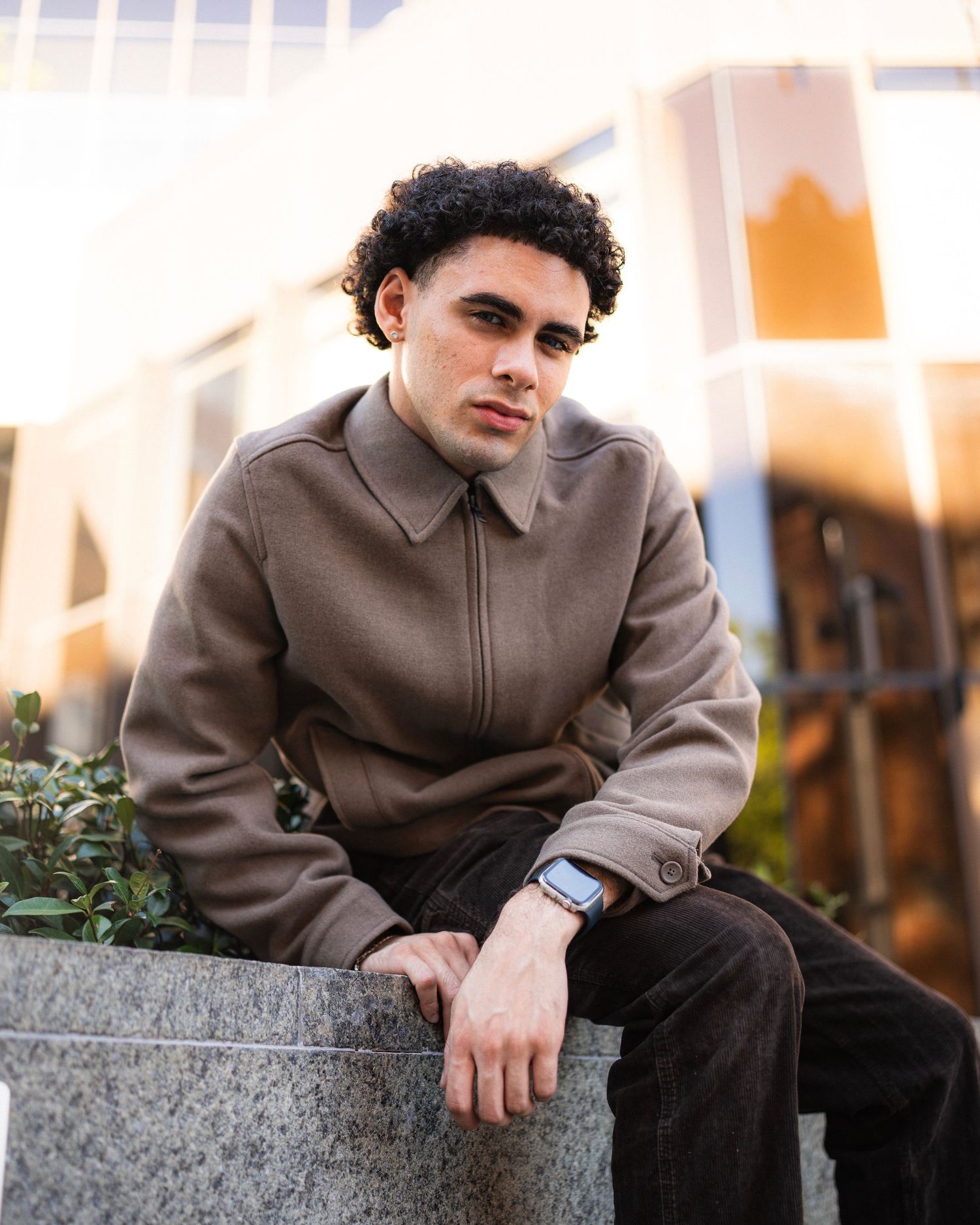 Casual portrait of a person in brown jacket sitting on concrete ledge during golden hour.